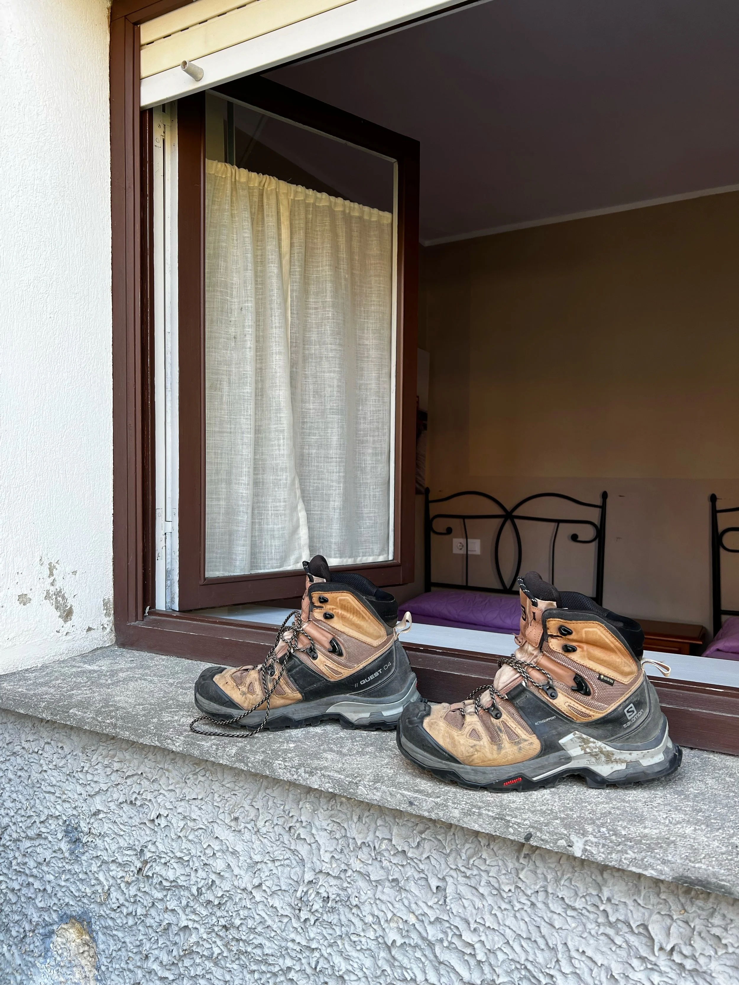 Pair of brown and black hiking boots on a concrete balcony ledge, with an open window and curtains in the background.