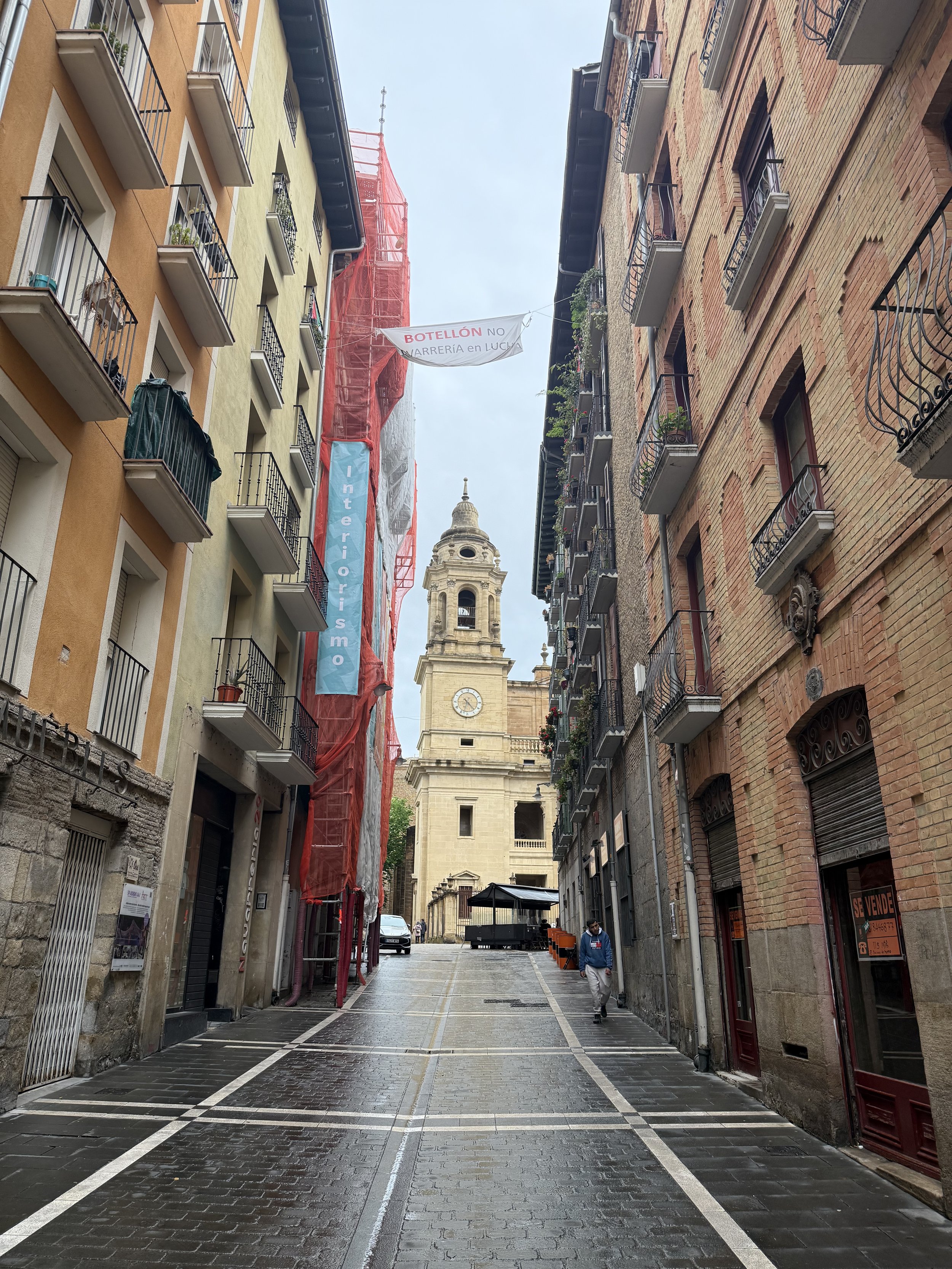 A narrow cobblestone street lined with multistory buildings on both sides, leading to a church with a clock tower in the background, with some people walking and signs hanging above the street.