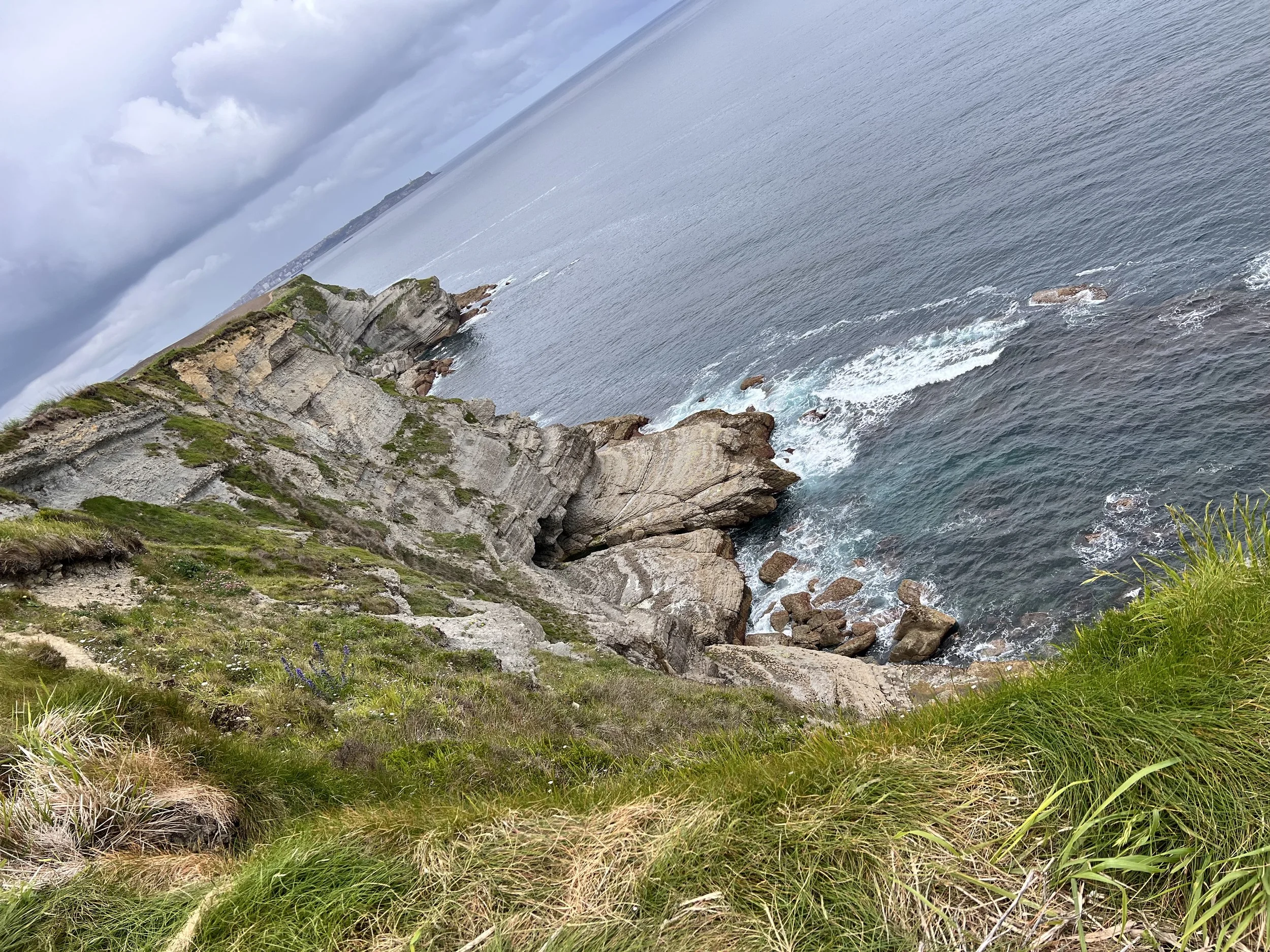 Rocky coastal cliffs with green grass and shrubs, overlooking the ocean with waves crashing against the rocks.