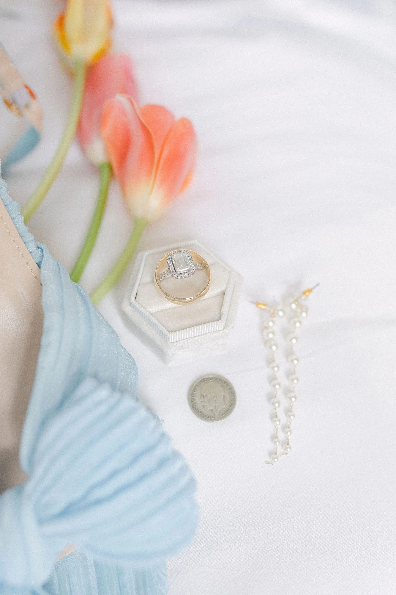 Close-up of jewelry including a diamond engagement ring in a white velvet ring box, pearl drop earrings, a quarter coin, and pink tulips on a white surface.