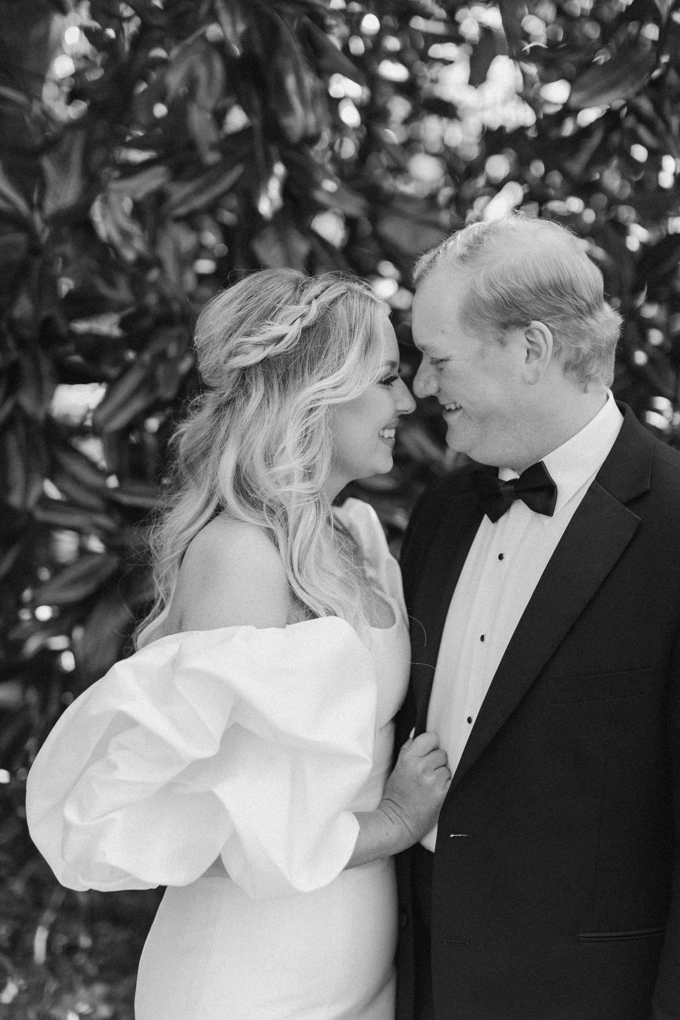 A bride and groom smiling and touching foreheads, standing close together outdoors with leafy background.