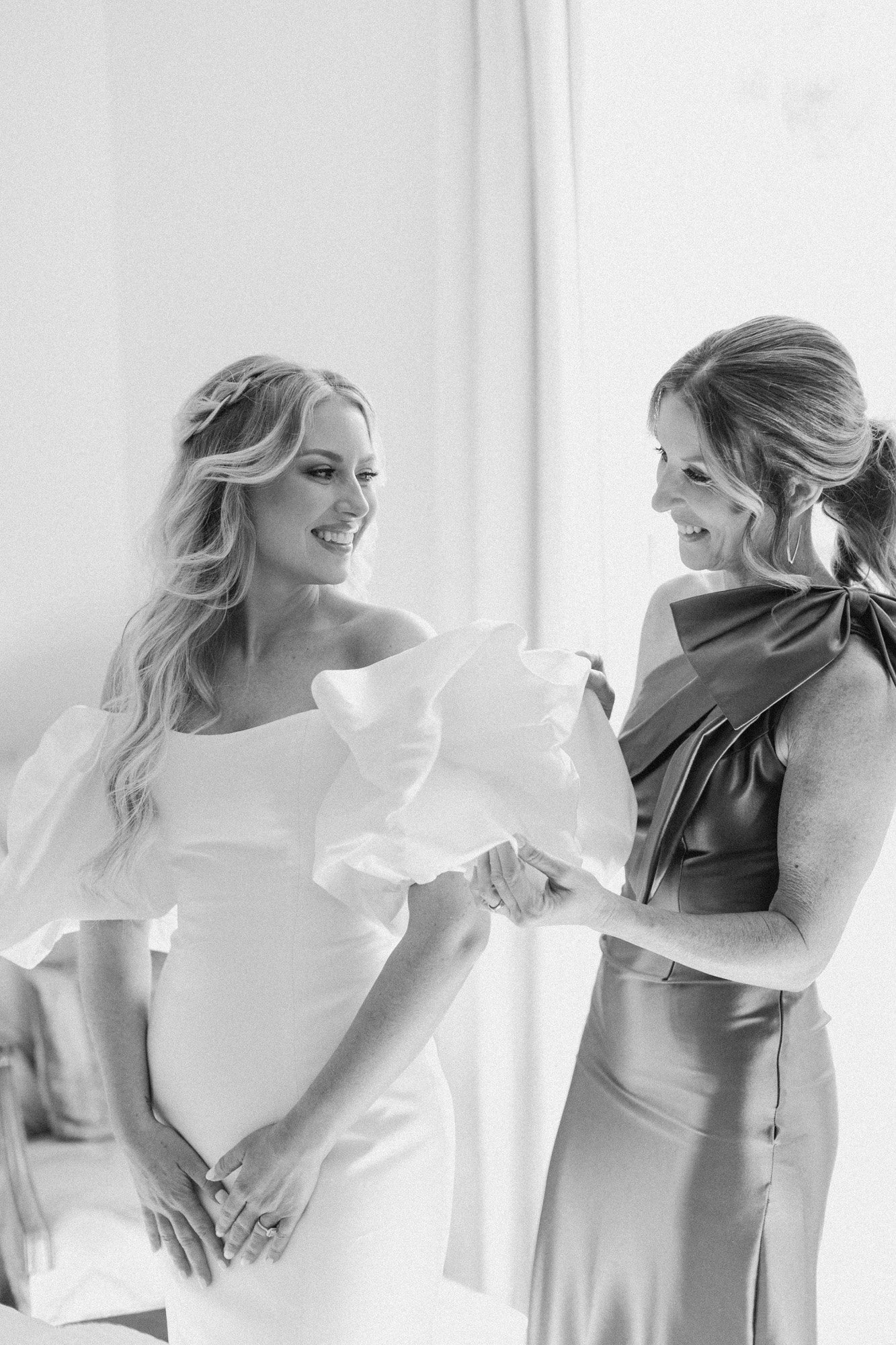 Two women, one in a wedding dress and the other in a satin dress with a bow, are smiling and exchanging a gift or veil.