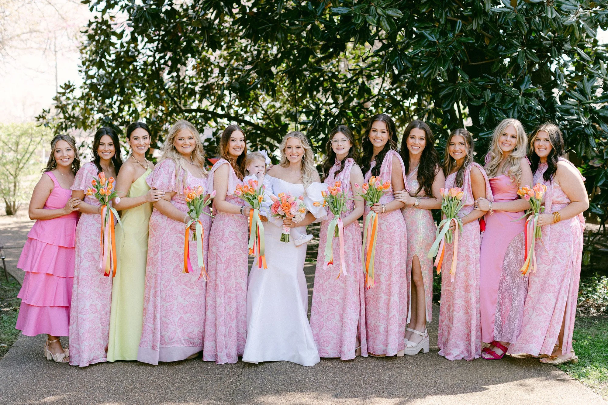 A bride and her bridesmaids posing outdoors in front of a large tree. The bride is wearing a white dress and holding a bouquet, surrounded by bridesmaids in pink and yellow dresses holding bouquets of orange tulips with ribbons.