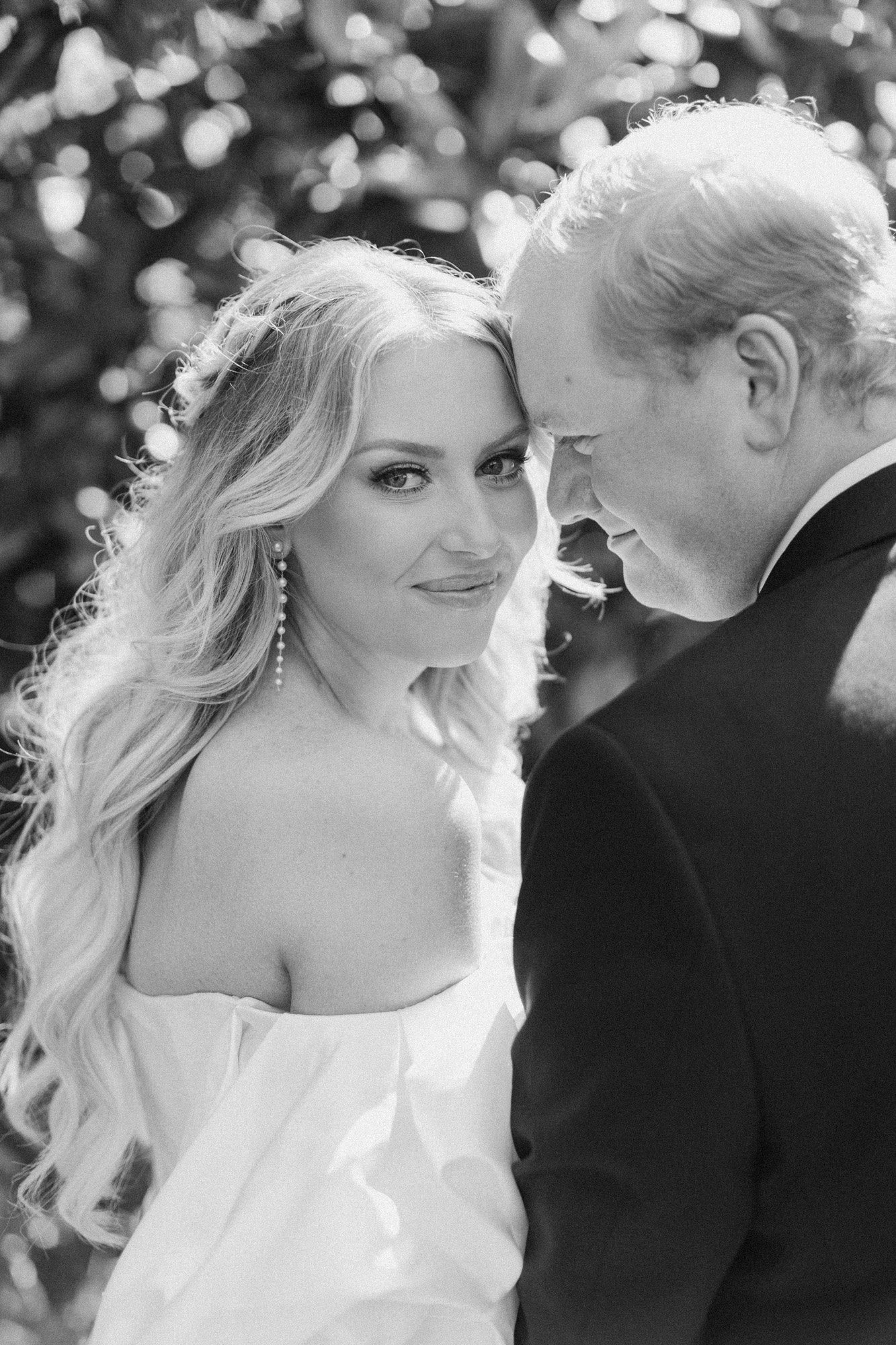 Black and white photo of a woman with long wavy hair and earrings looking at camera, standing close to a man with light-colored hair, both dressed formally, outdoors with blurred foliage in background.