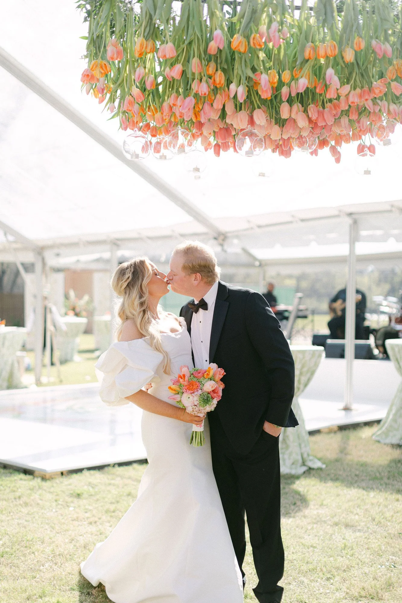 Bride and groom kissing outdoors at their wedding under a floral canopy, with a musician playing guitar in the background.