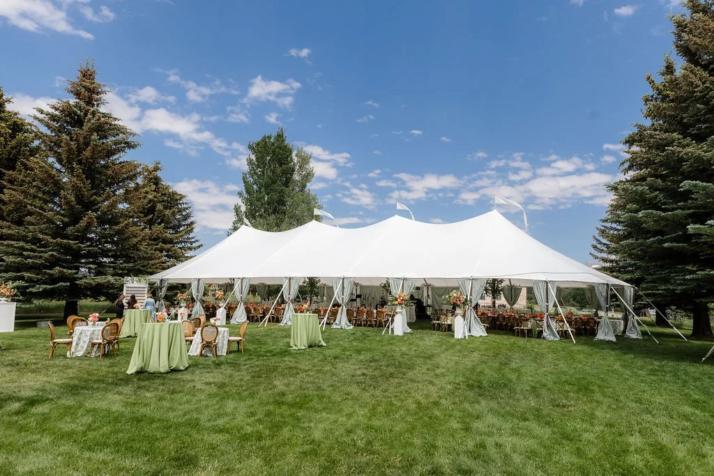 Large white outdoor event tent set up on a grassy field with tables and chairs inside. Small tables with green tablecloths are outside, and tall trees surround the area under a partly cloudy blue sky.