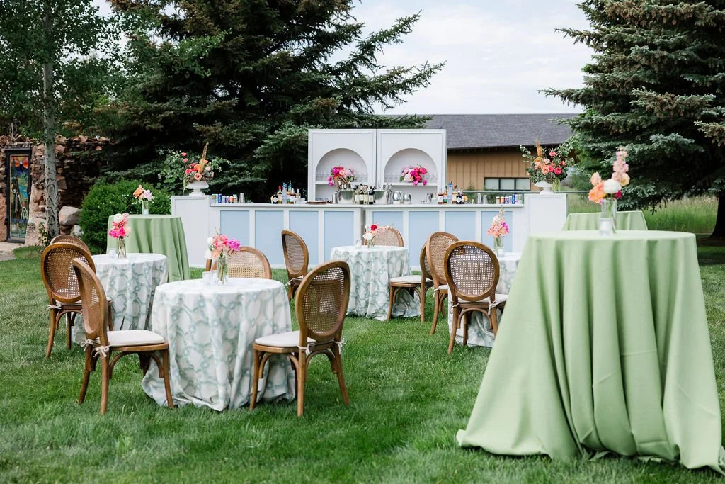 Outdoor event setup with round and high-top tables, decorated with pink floral arrangements, a bar area with drinks and flowers, surrounded by trees and a building in the background.