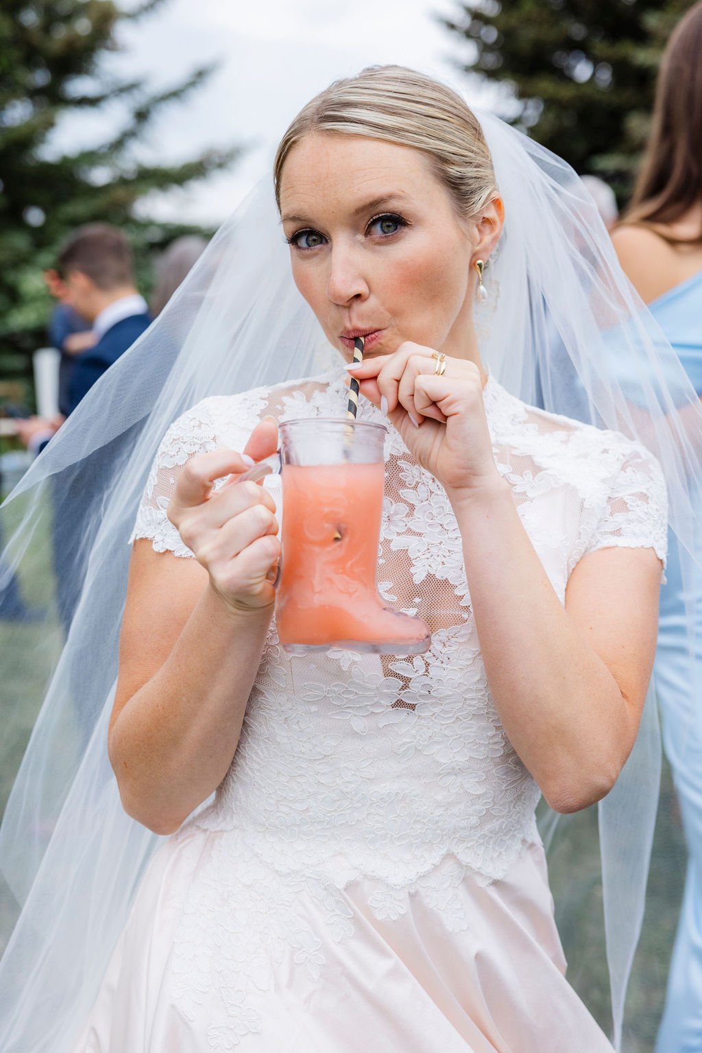 Bride in wedding dress and veil drinking pink drinks through a black and white striped straw at an outdoor wedding.
