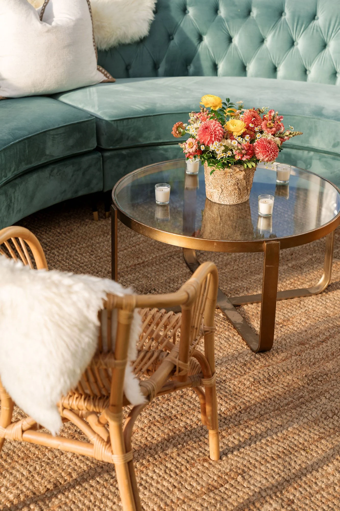 A living room with a round glass coffee table with a flower arrangement, surrounded by a green velvet tufted sofa and a rattan chair with a white fluffy throw.