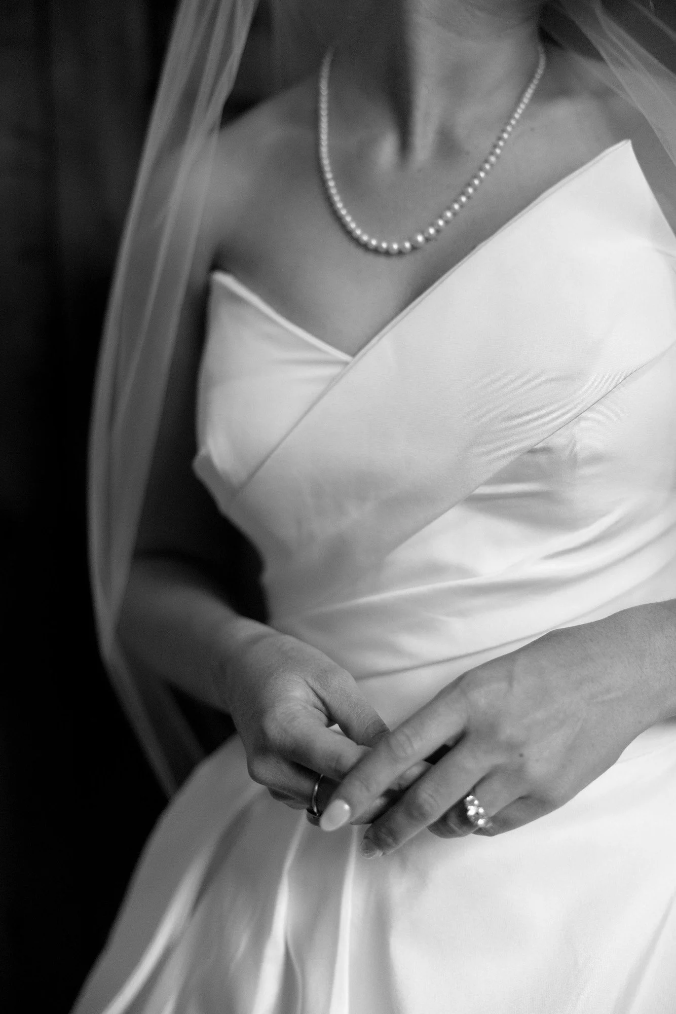 Black and white photo of a bride wearing a strapless satin wedding dress, pearl necklace, and veil, with hands clasped at her waist, showing wedding rings.