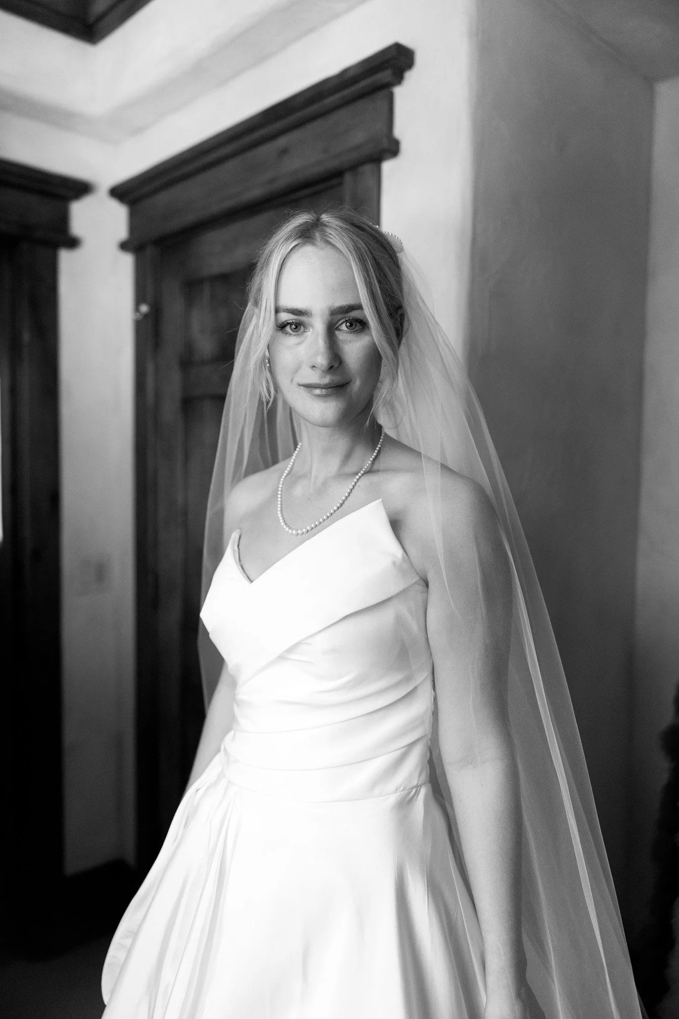 Black and white photo of a young woman in a wedding dress and veil, standing indoors in front of wooden doors, looking at the camera.