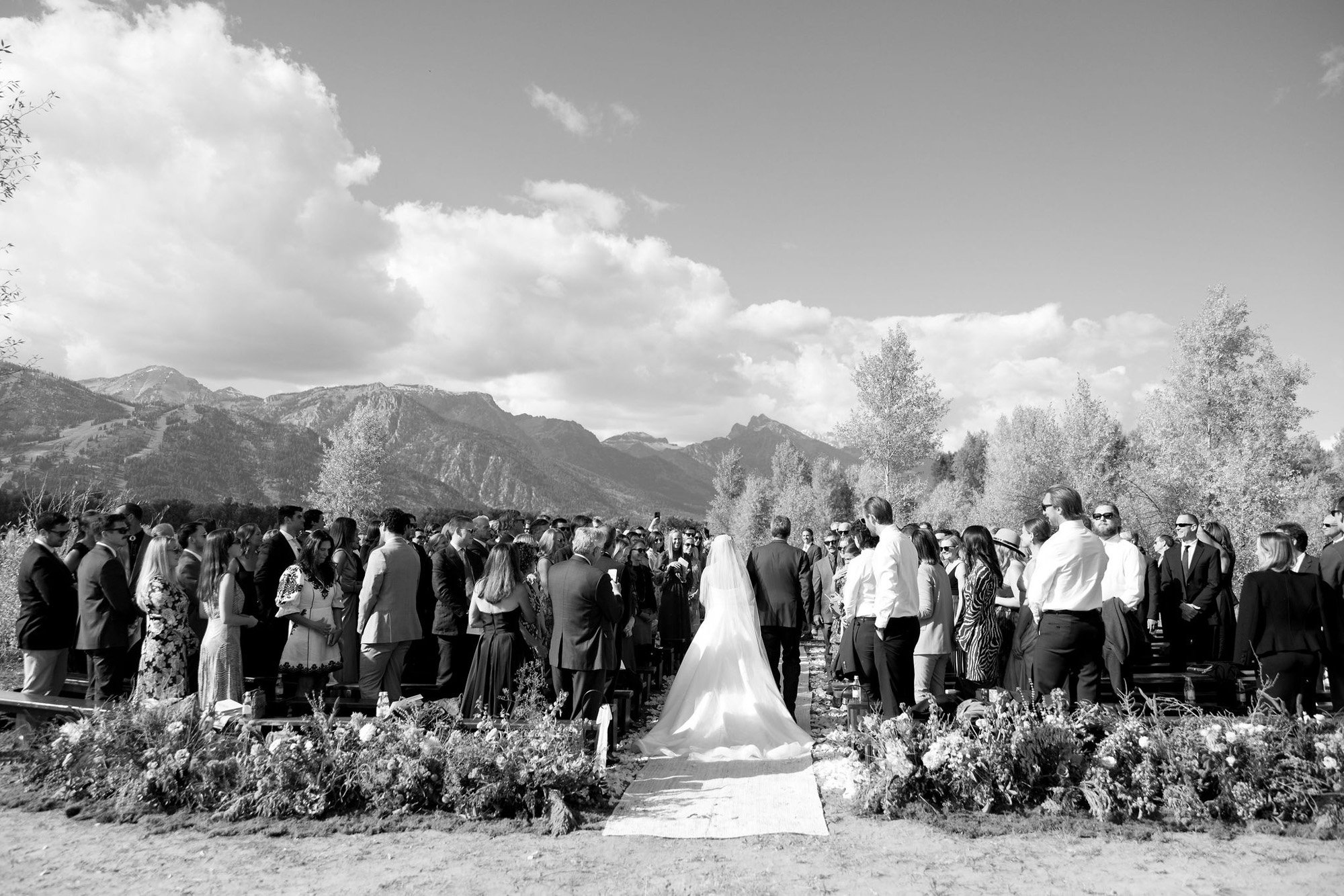 A black and white photo of an outdoor wedding ceremony with guests standing on either side of an aisle, a bride in a long white dress and veil facing the officiant, mountains and sky in the background.