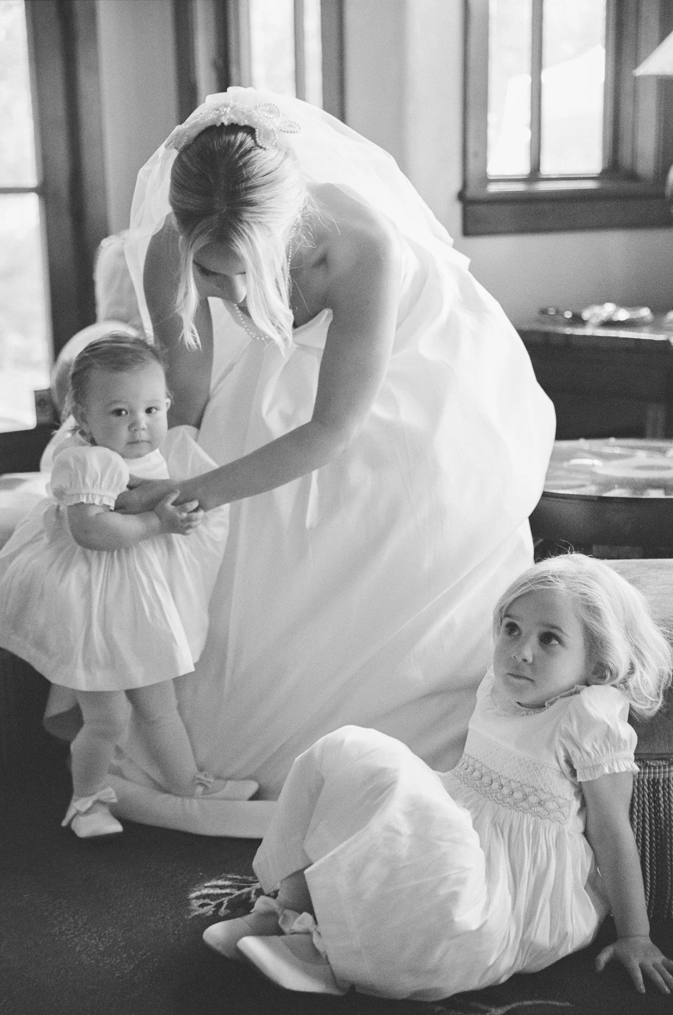 A bride in a wedding dress helping two young girls into their dresses in a room with windows.