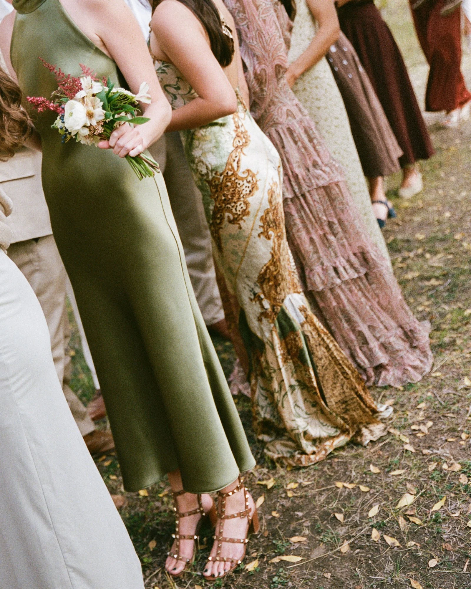 Line of women standing outdoors, dressed in elegant attire, with one holding a bouquet of flowers.