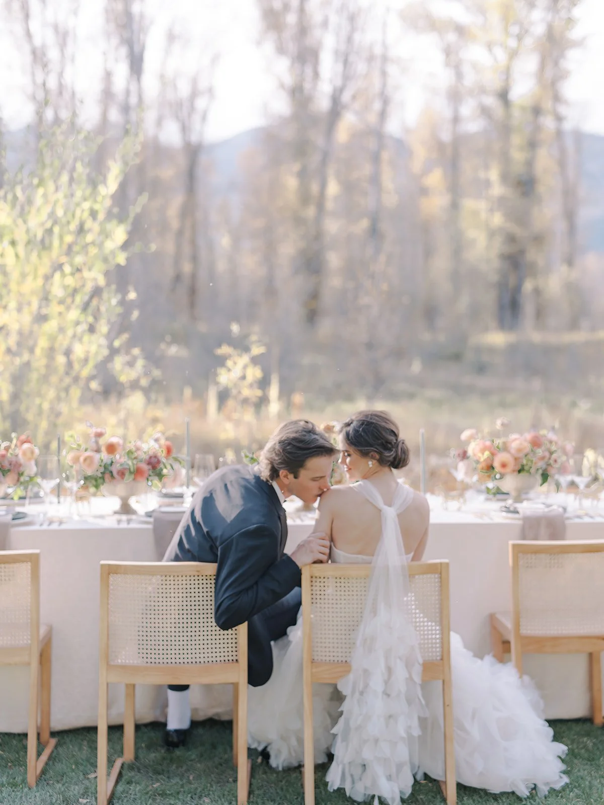 A couple dressed in wedding attire sitting outside at a decorated wedding reception table with flowers, with a fall landscape in the background.