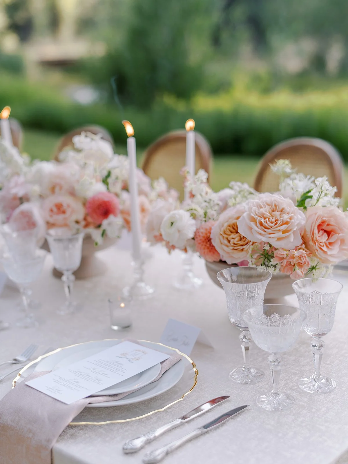 Elegant outdoor table setting with pink and white flowers, glassware, candles, and a menu on a white tablecloth, overlooking a green landscape.