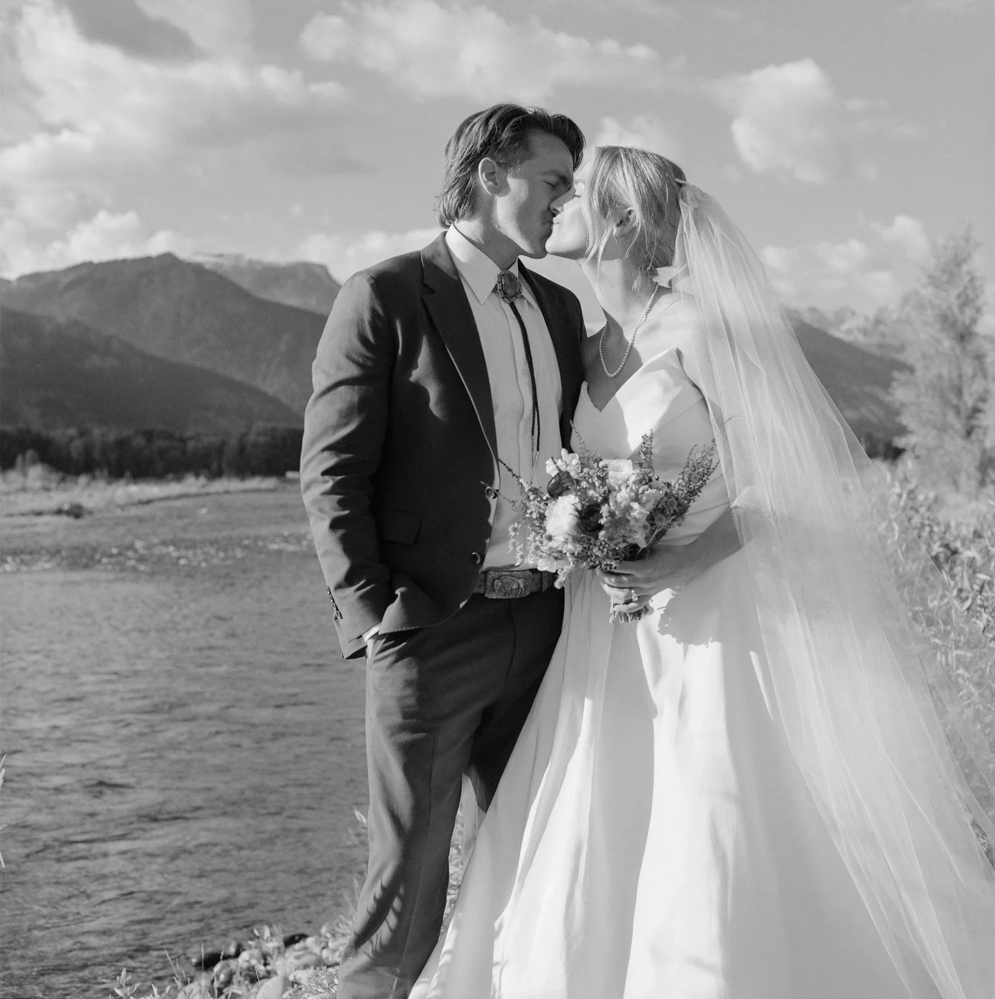 Black and white photo of a bride and groom sharing a kiss outdoors near a body of water with mountains in the background. The bride is wearing a wedding dress and veil, holding a bouquet, and the groom is in a suit with hands in his pockets.