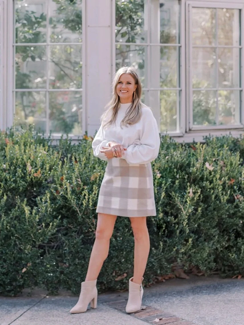 A woman standing outdoors in front of a large glass window, wearing a white long-sleeve blouse, beige plaid skirt, and beige ankle boots, smiling at the camera.