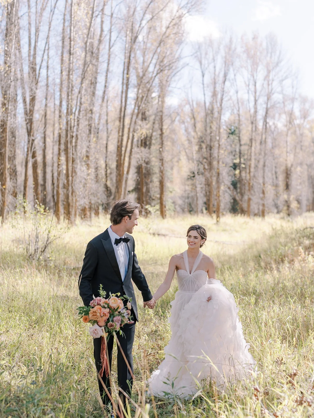 Bride in a lace gown and groom in a tuxedo walk hand in hand near a pond during their Jackson Hole wedding.