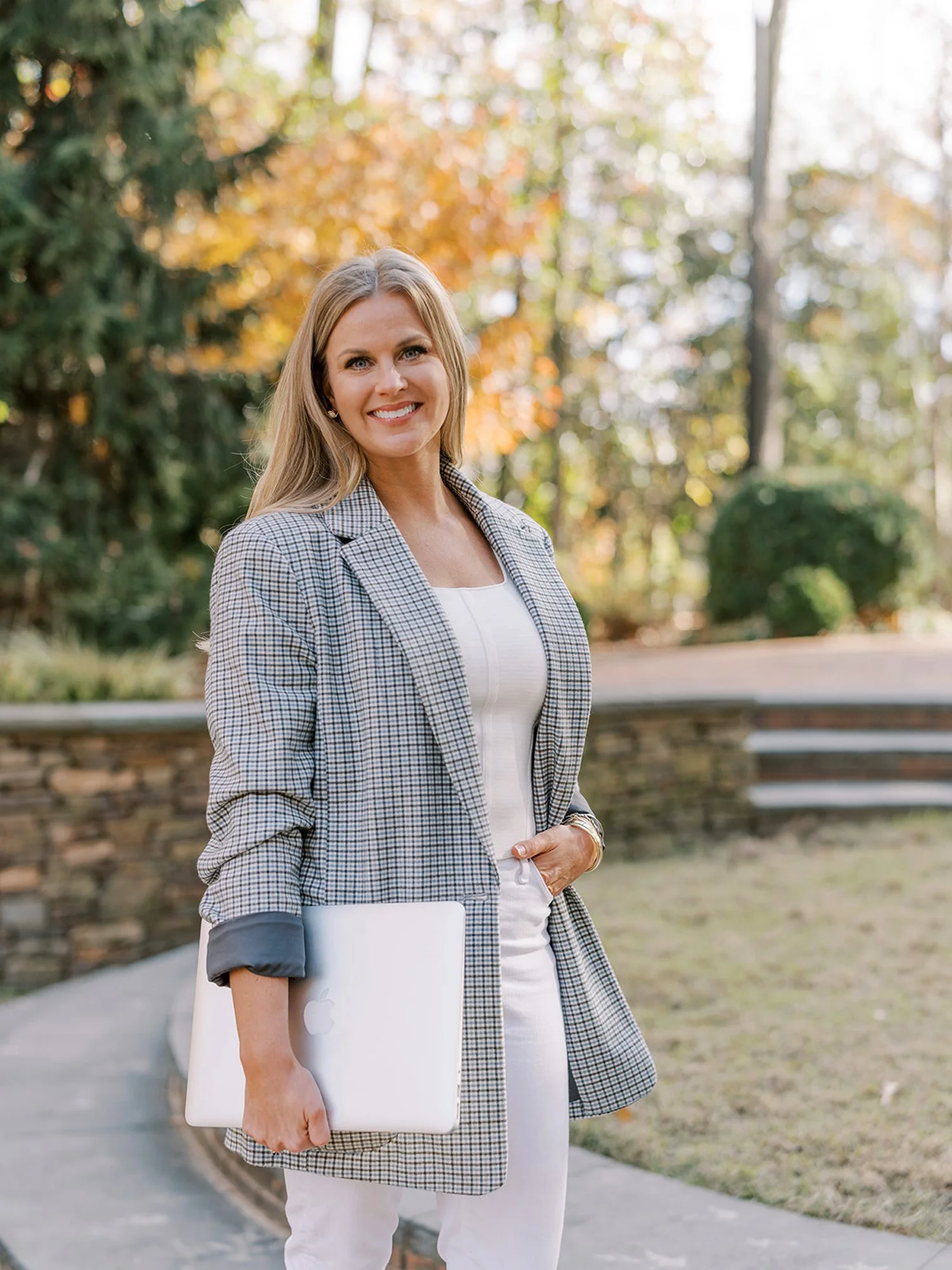 A woman smiling outdoors holding a closed silver MacBook laptop, wearing a checked blazer over a white top, with an autumnal background of trees and a stone wall.