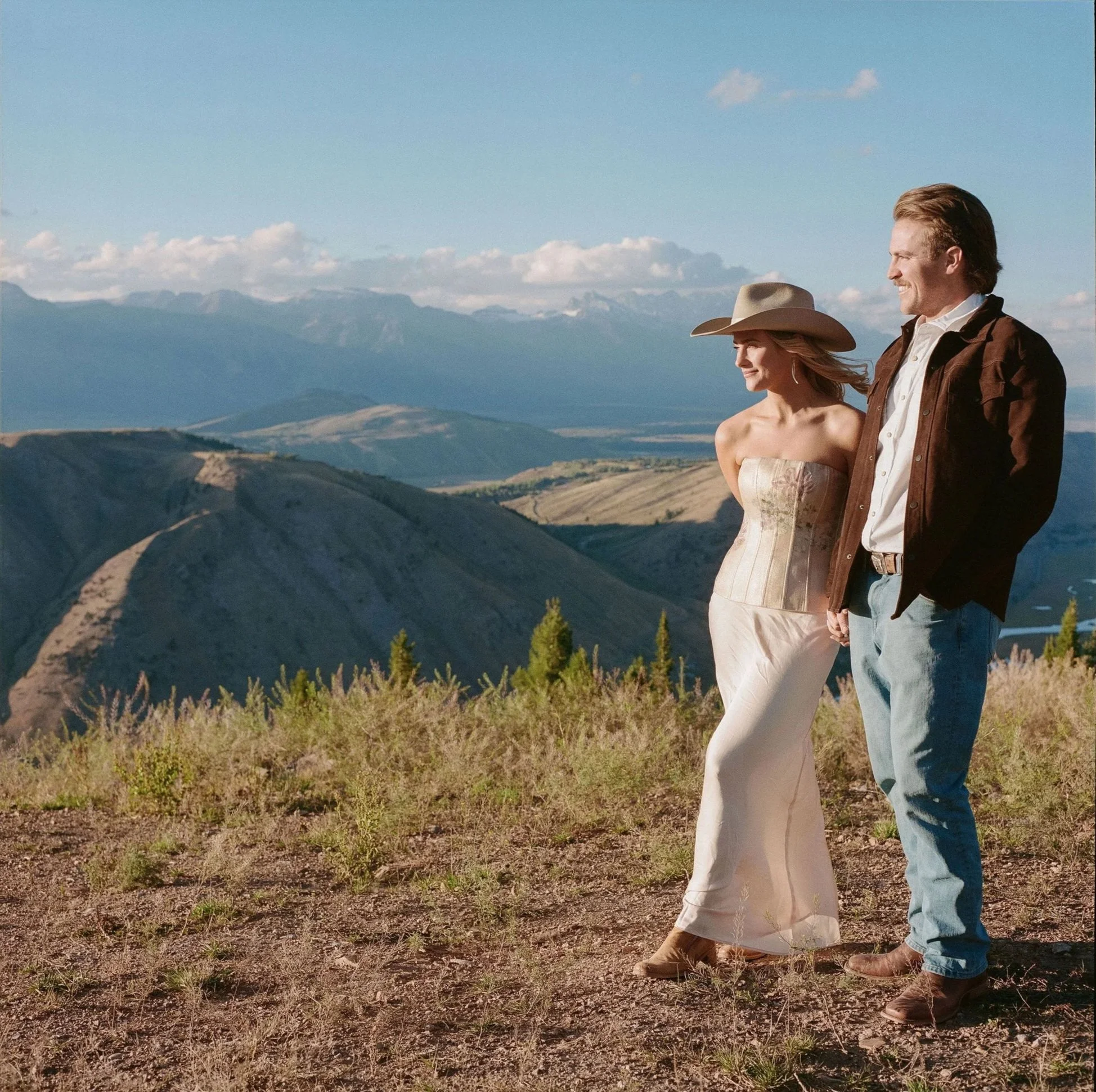A man and woman standing on a hilltop with mountains in the background, enjoying a sunny day. The woman is wearing a wide-brimmed hat and a strapless dress, and the man is dressed in a jacket and jeans.