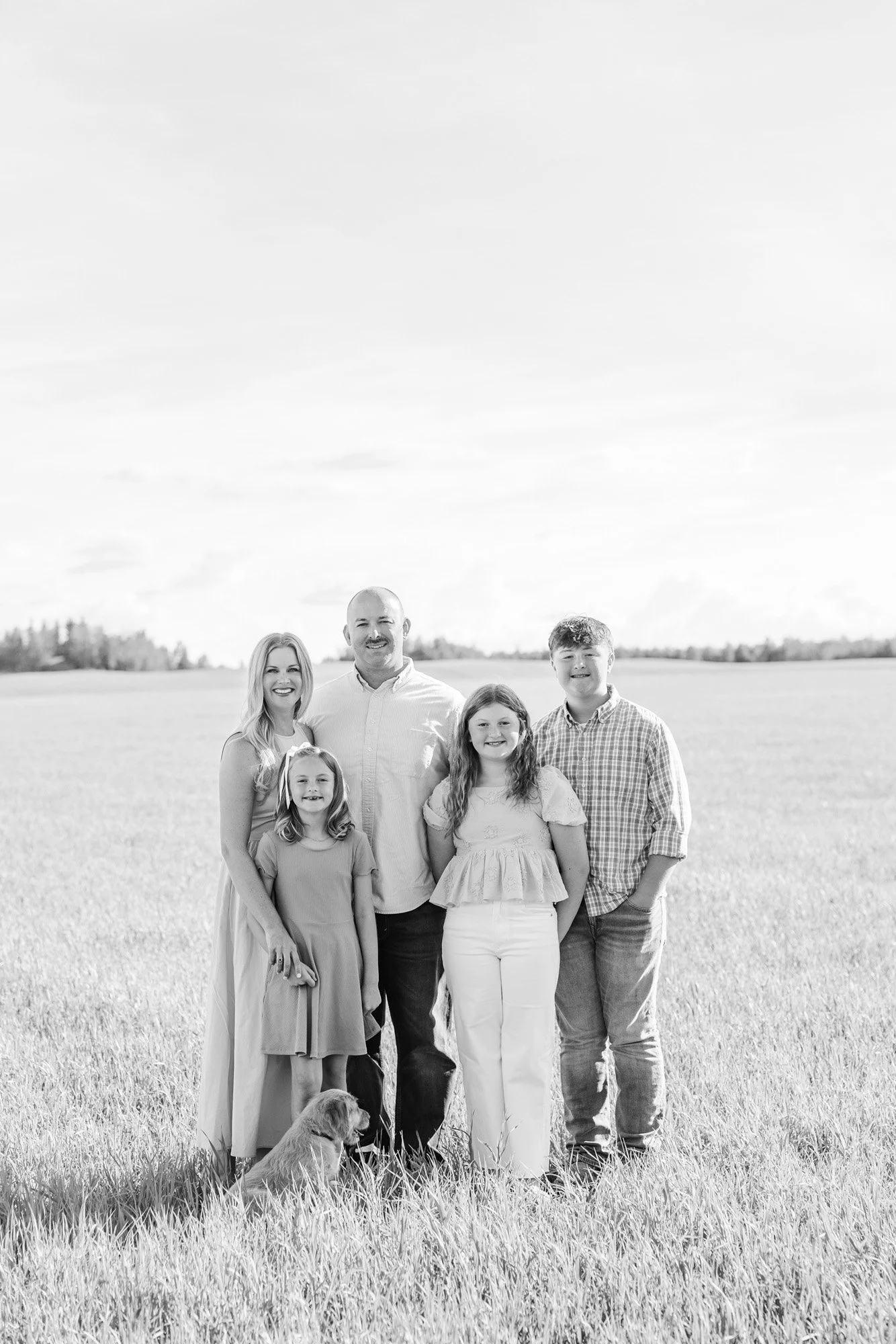 Black and white photo of a five-person family standing outdoors in a grassy field with a dog. The family includes two adults and three children, all smiling and dressed casually.