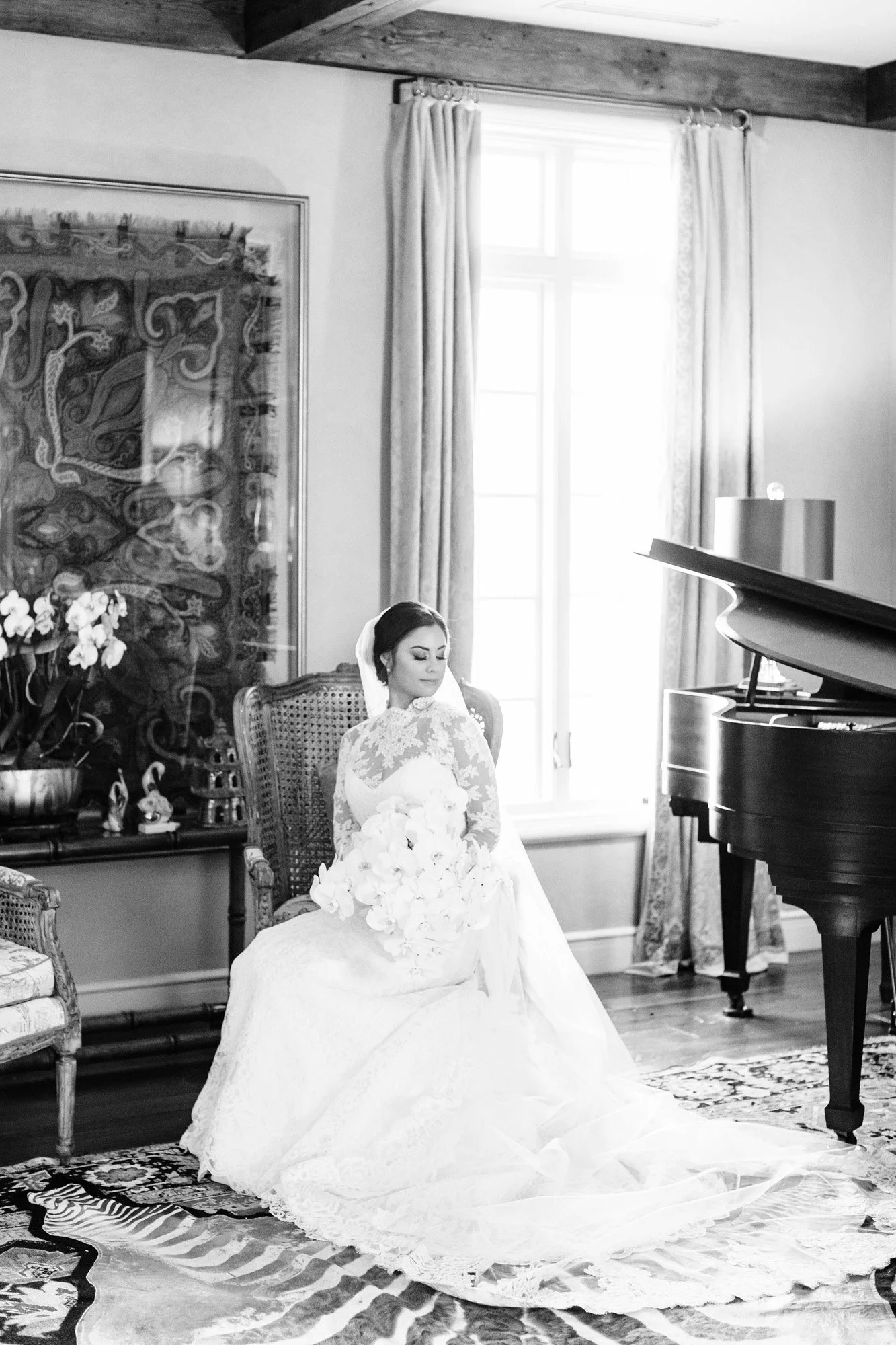 A bride sitting on a vintage chair in a well-lit, elegant room, holding a bouquet of flowers, with a grand piano nearby, in black and white.