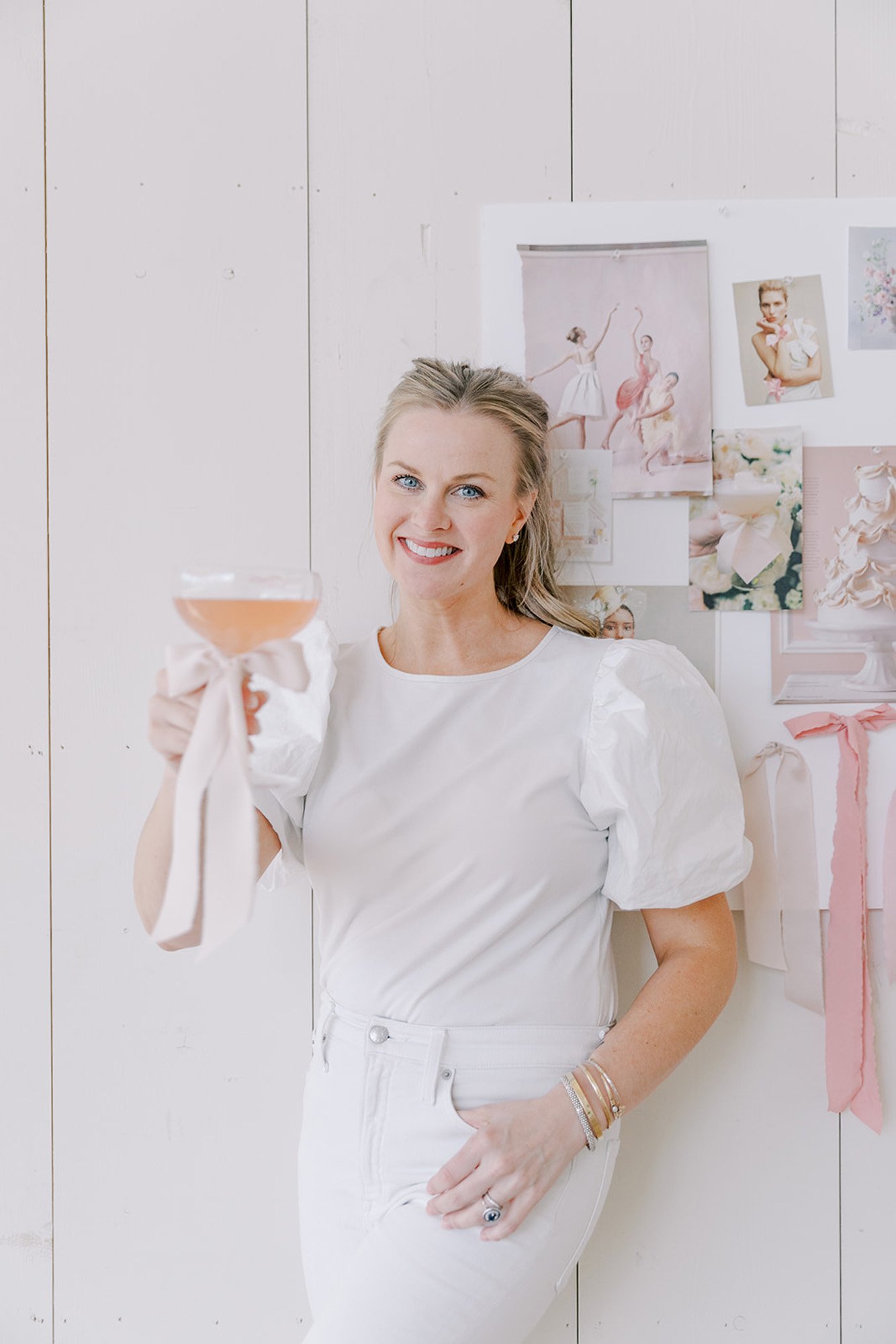 A woman holding a pink cocktail with a pink ribbon in a casual white outfit, standing in front of a white wall with floral and ballet-themed pictures.