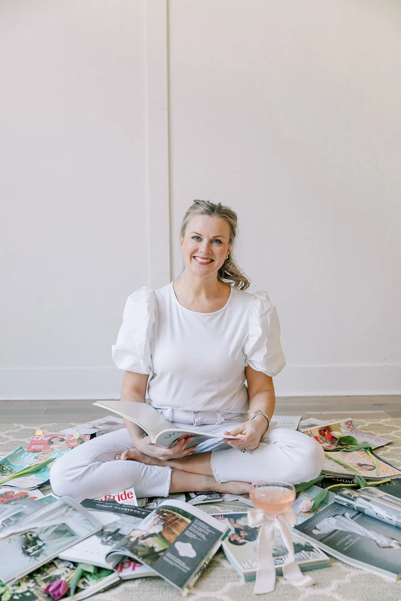 A woman sits cross-legged on the floor, surrounded by magazines and books, holding an open magazine and smiling at the camera.