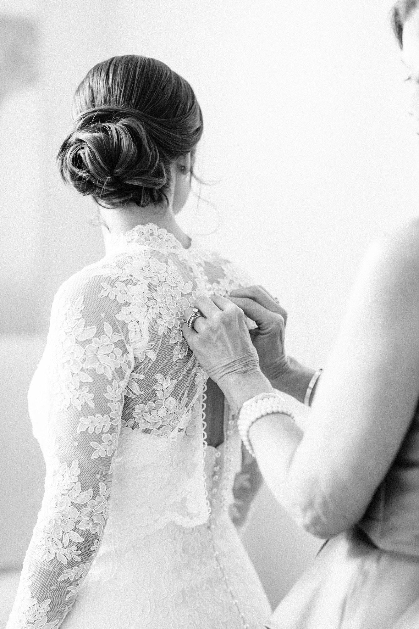 A bride getting ready, with someone adjusting her lace wedding dress.