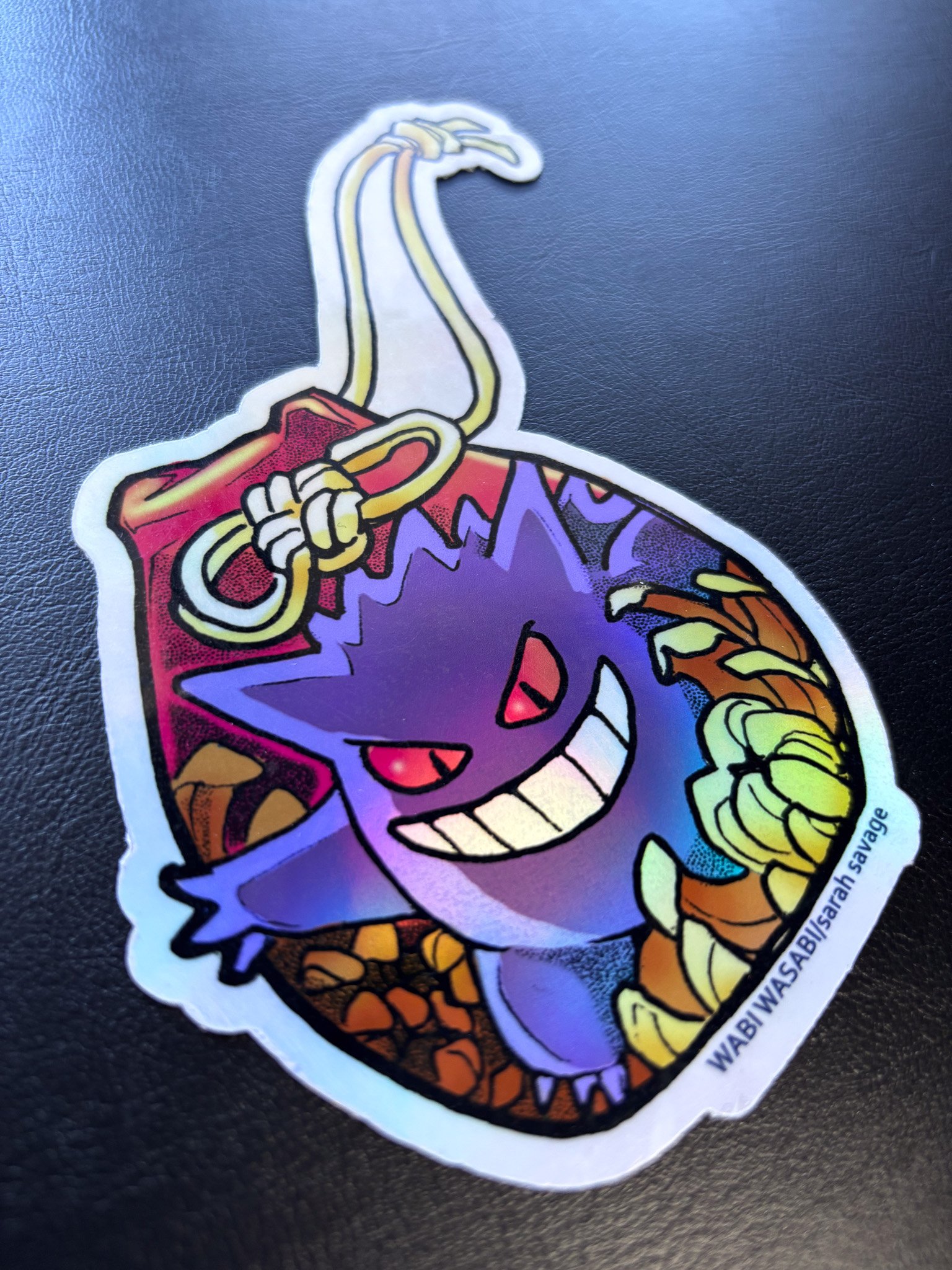 Holographic sticker of a purple, mischievous creature with red eyes, big grin, and a large toothy smile, holding a gold chain with a ring, with a background of rocks or soil.