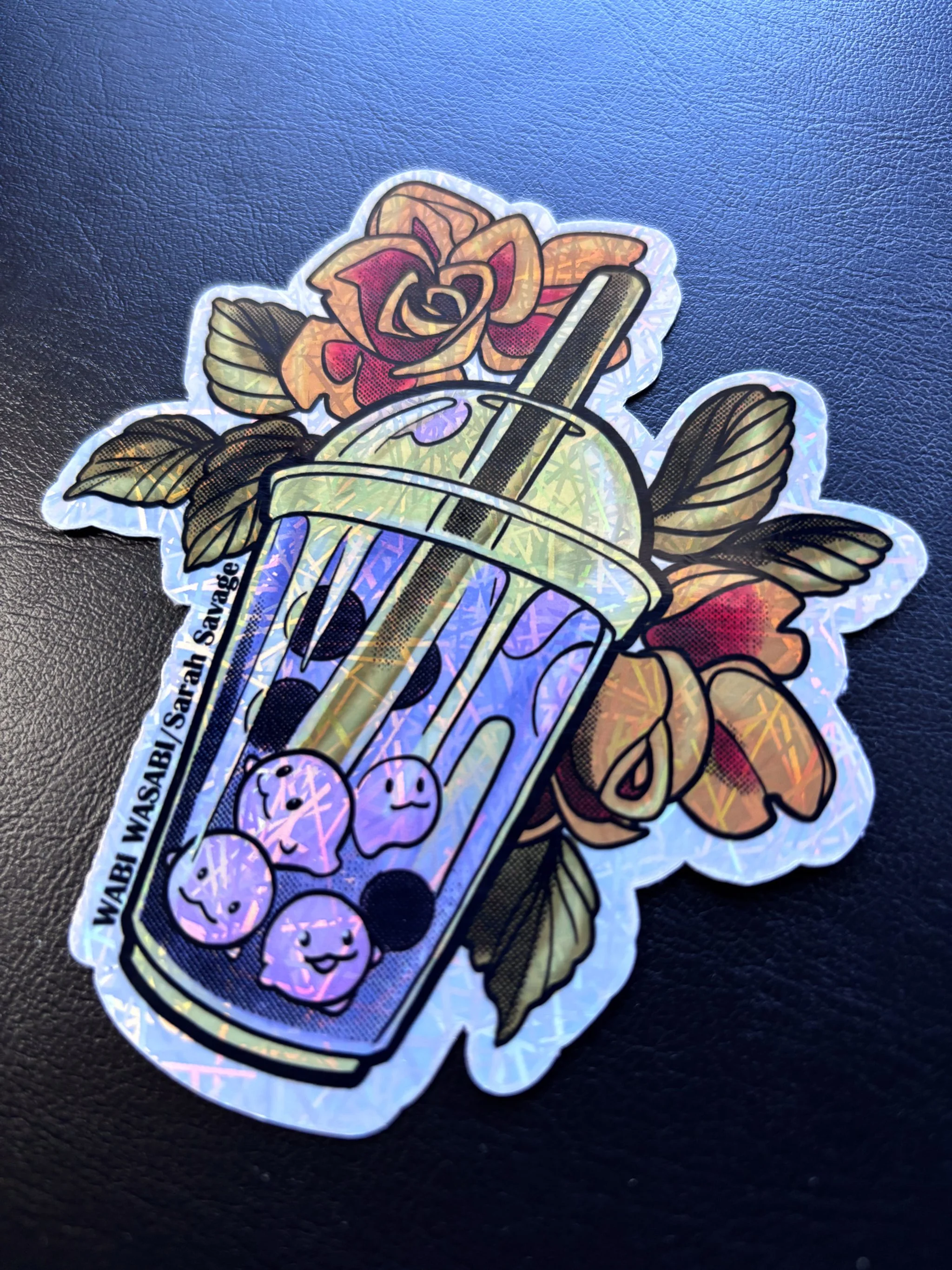 Holographic sticker of bubble tea with tapioca pearls and smiling faces, surrounded by orange roses and green leaves.