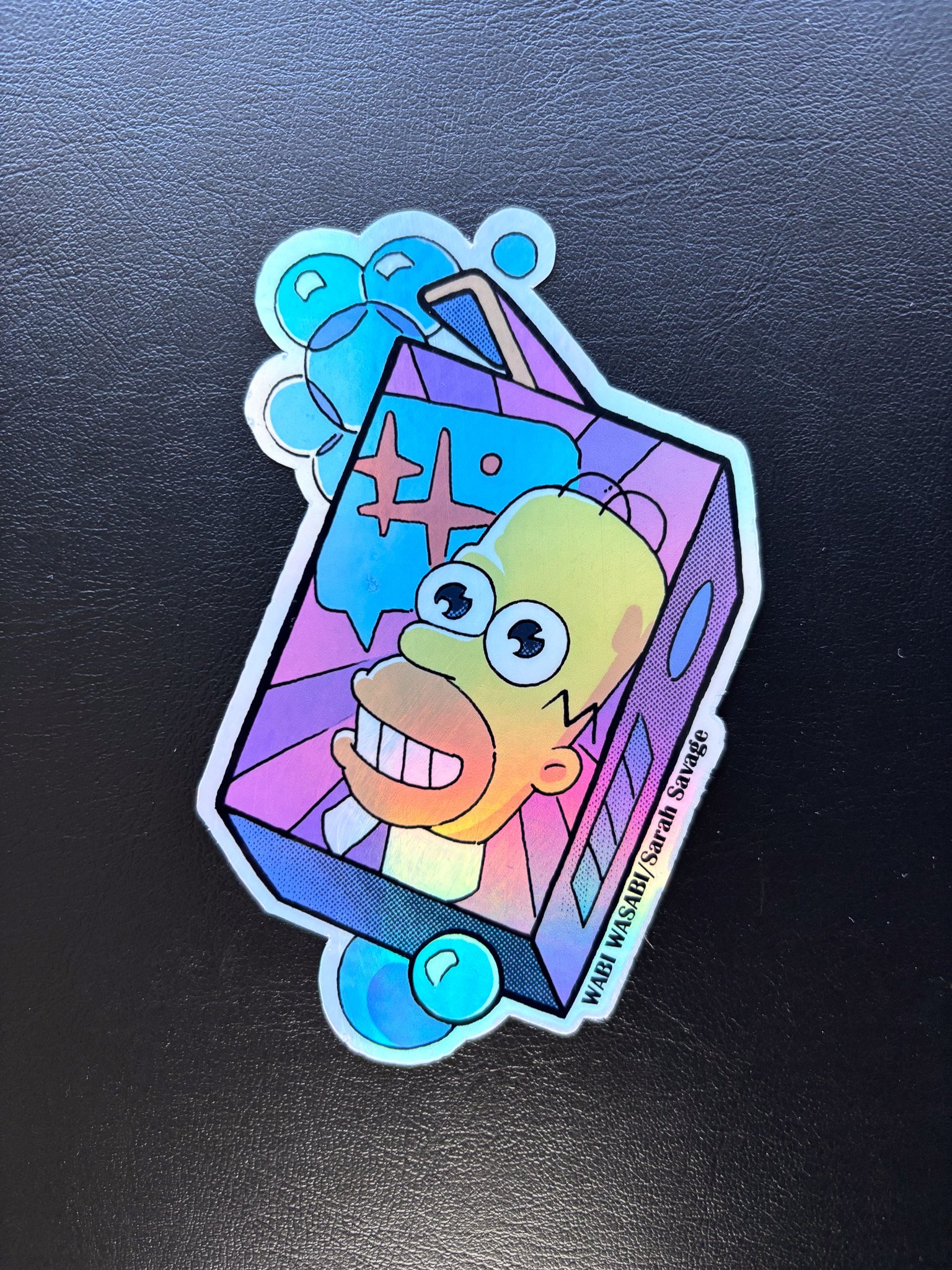 Sticker of a cartoon Homer Simpson with wide eyes and a big grin, inside a colorful box with bubbles and abstract shapes, on a black background.