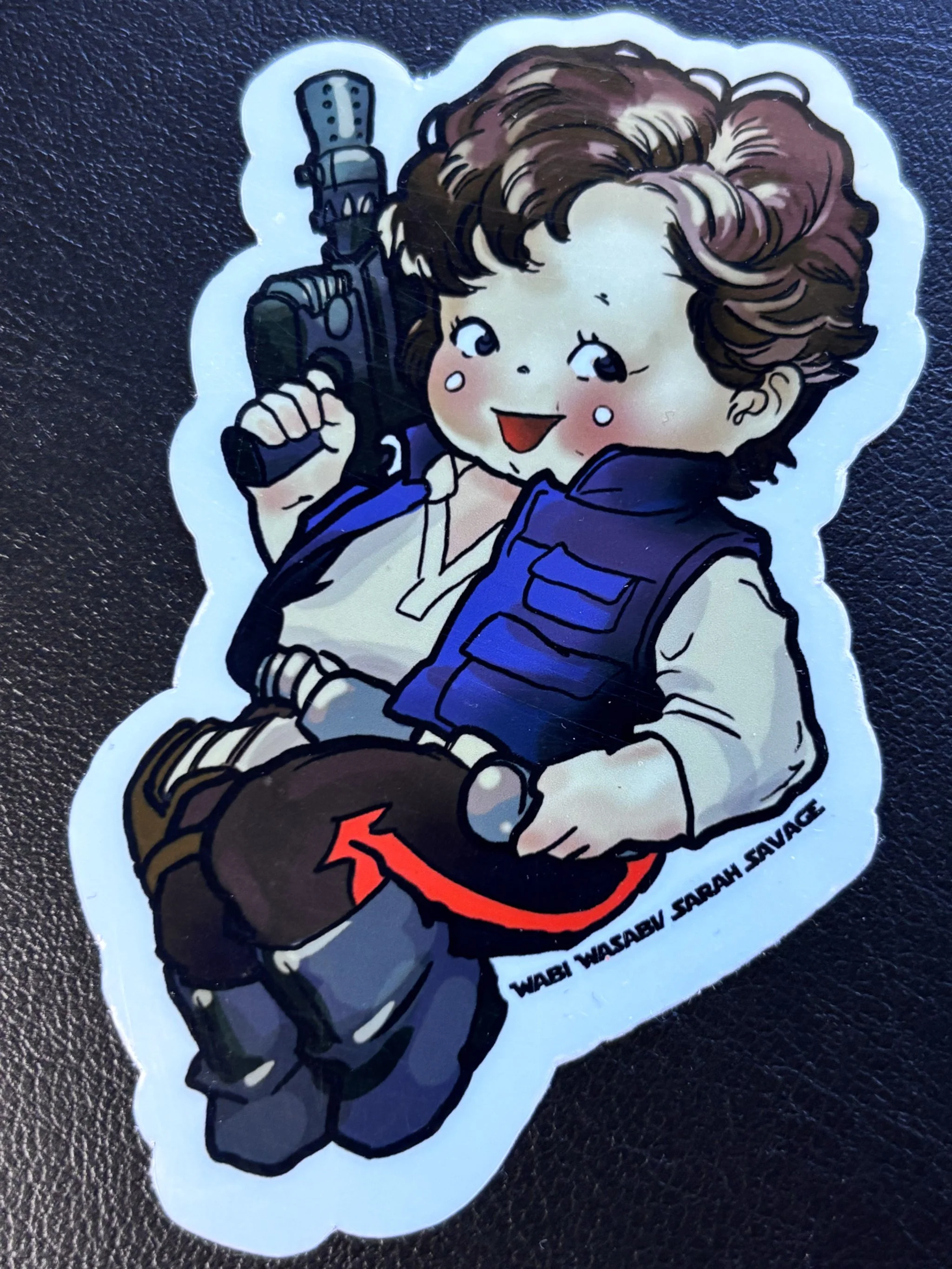 Sticker of a cartoon girl with brown hair, holding a gun, wearing a vest and boots, sitting with her legs crossed, smiling. The text on the sticker reads 'WABI WASABI SARAH SAVAGE'.