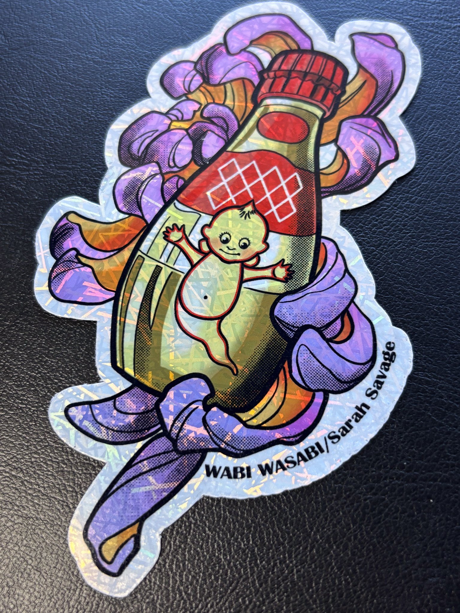 Holographic sticker of a baby riding a jalapeño pepper surrounded by purple and orange flames with the text "WABI WABY/Sarah Savage" at the bottom.