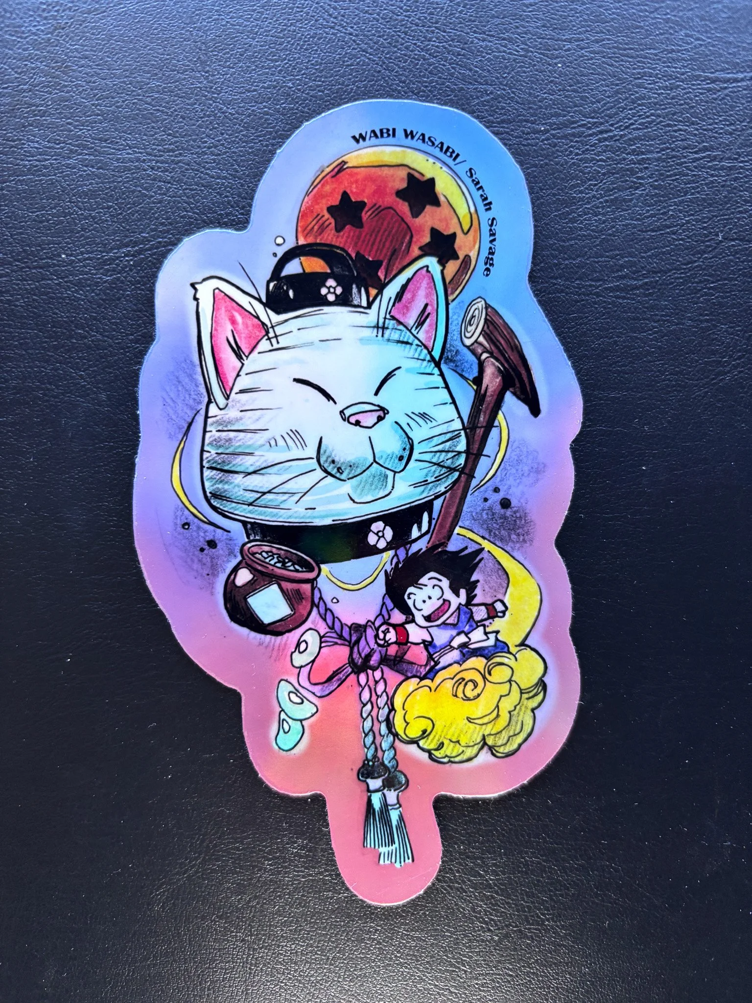 Colorful sticker depicting a smiling cat wearing a collar with a small jar and a broomstick, a young person with black spiky hair riding a yellow cloud, a full moon with black stars, and the text 'WABI WASABI Sataran Savage' at the top.