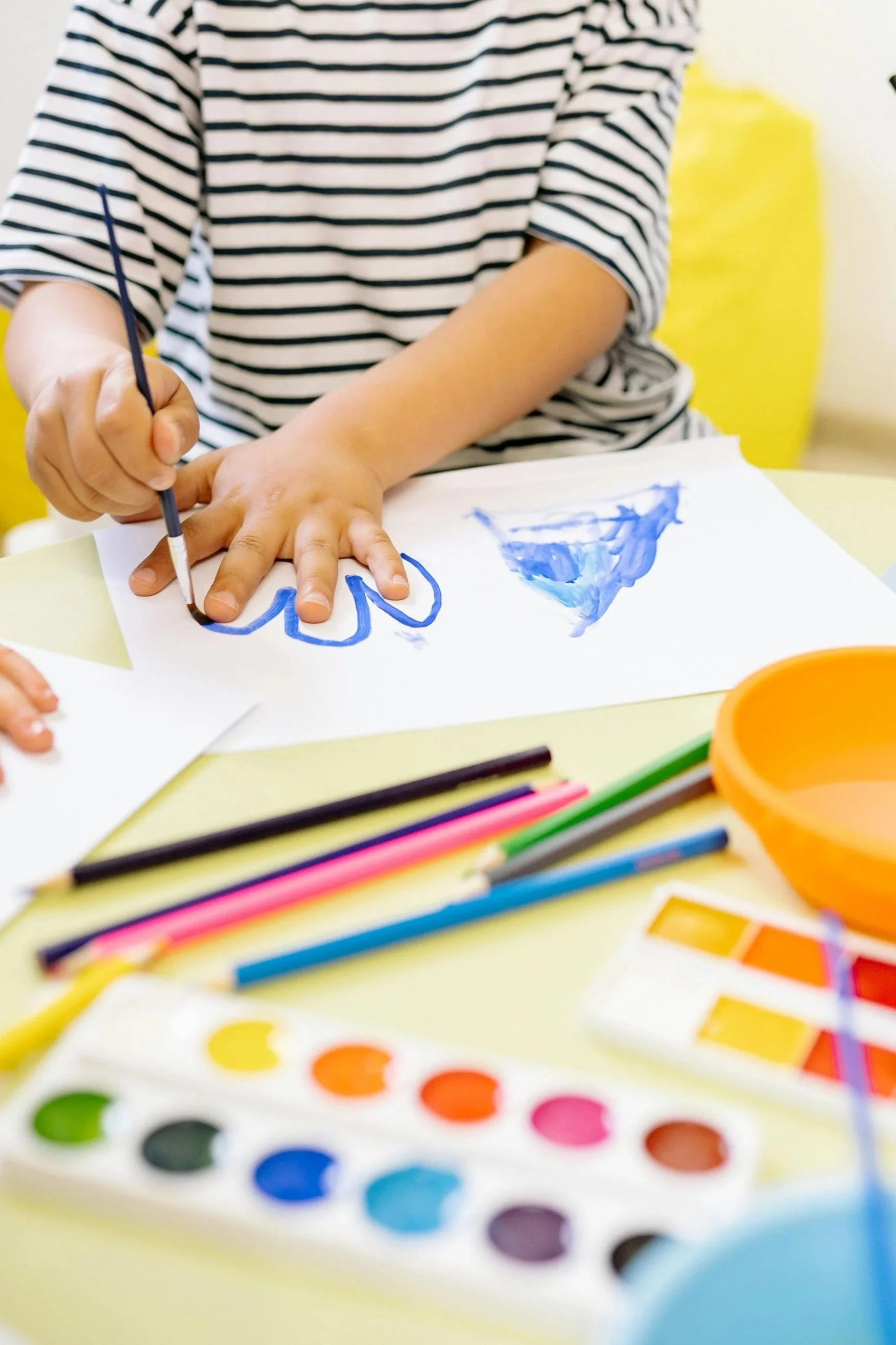 Child wearing a striped shirt painting with watercolor paints and colored pencils on paper, with art supplies on a table.