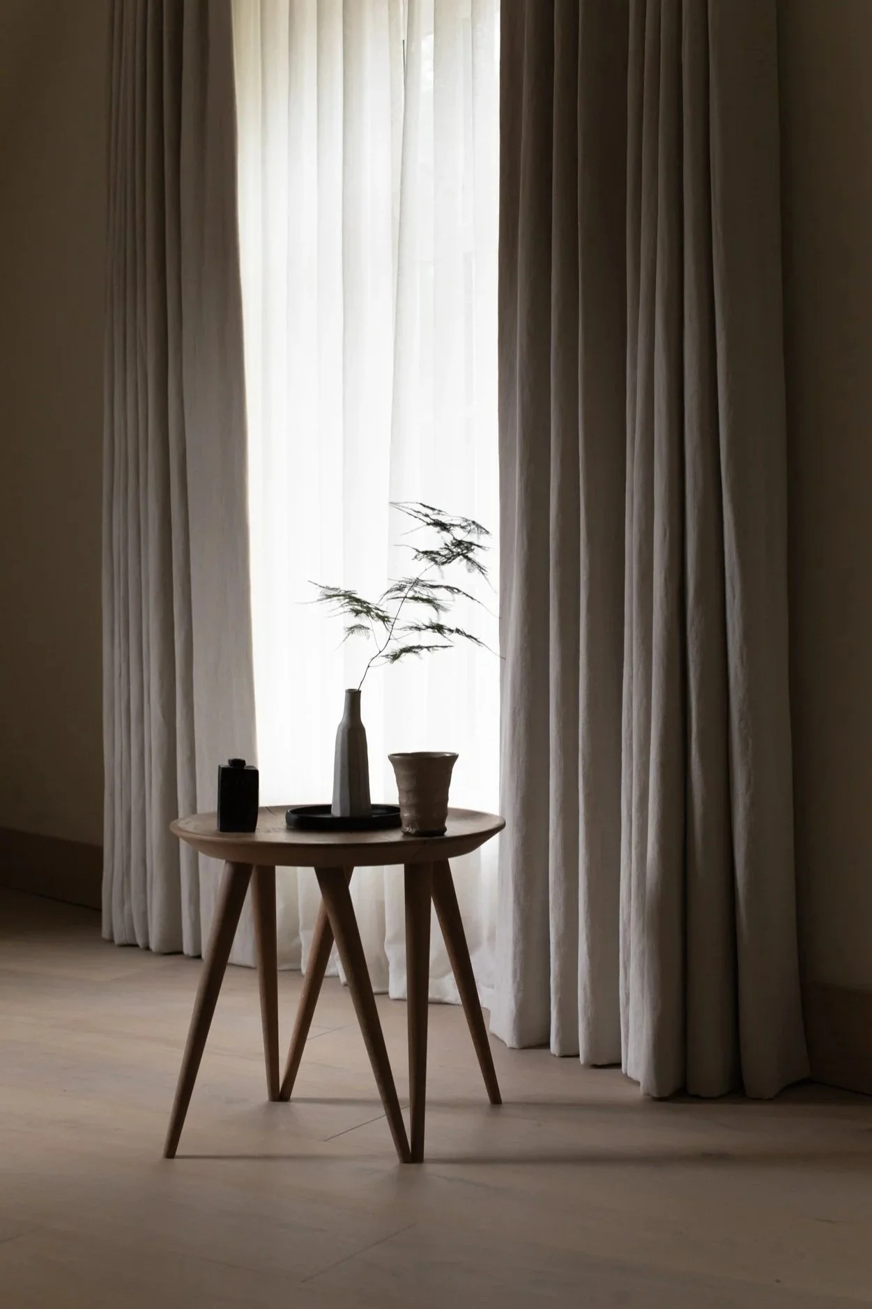 A small wooden table with a pottery vase containing a single branch with leaves, a small container, and a tray, set in front of sheer curtains.