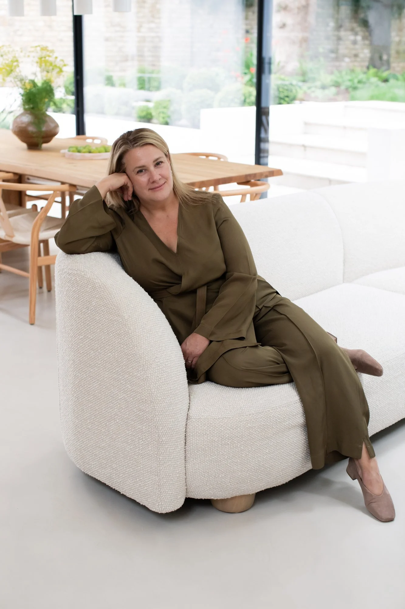 A woman in olive green pajamas sitting sideways on a white couch in a modern living room with large windows and a wooden dining table in the background.