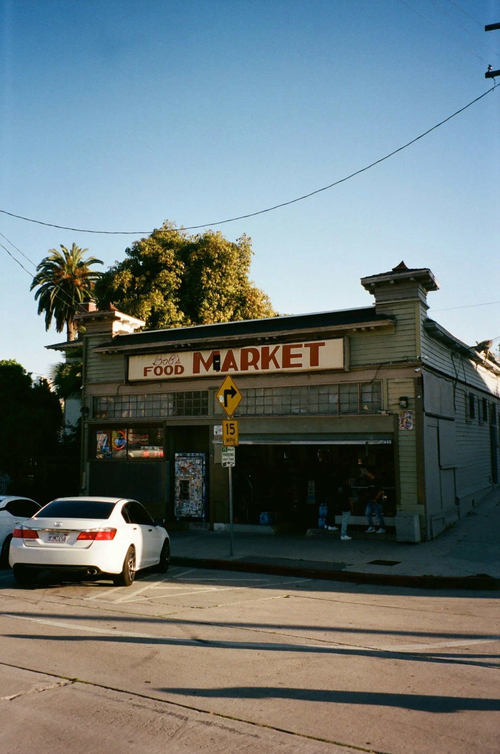 An old wooden building with a sign that reads 'Bob's Food Market'. The building has a vintage look with large windows and a garage-style door. There are a few cars parked in front, including a white sedan. A yellow traffic sign indicating a right curve and a 15 MPH speed limit is visible. The background includes trees and a clear blue sky.