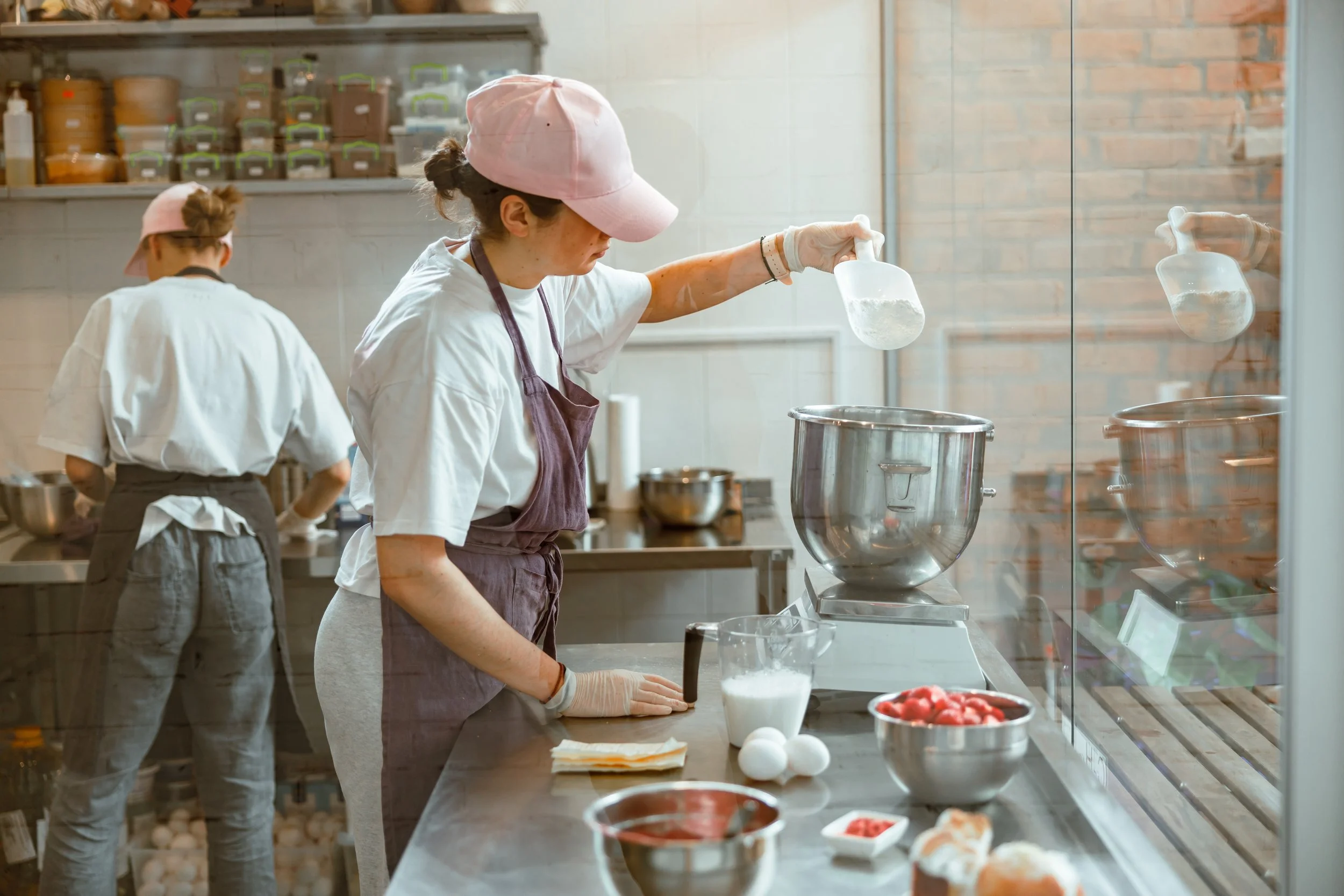Two women in a commercial kitchen, one in the foreground pouring ingredients into a large mixing bowl, the other in the background preparing food, with various ingredients and tools on the stainless steel countertop.