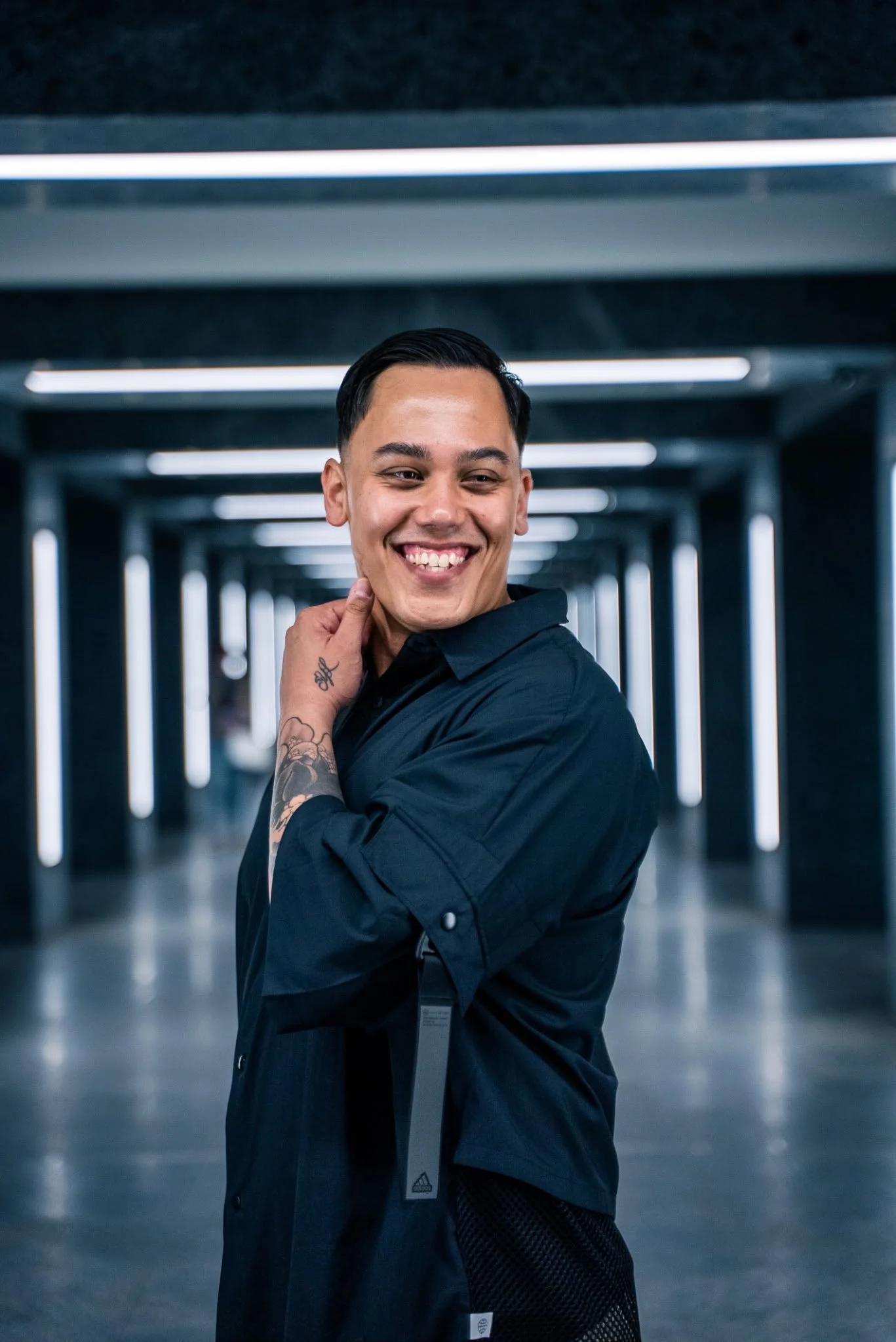 A smiling man with short black hair, tattoos on his left arm, wearing a dark blue shirt, standing in a modern, illuminated corridor with geometric ceiling lights.