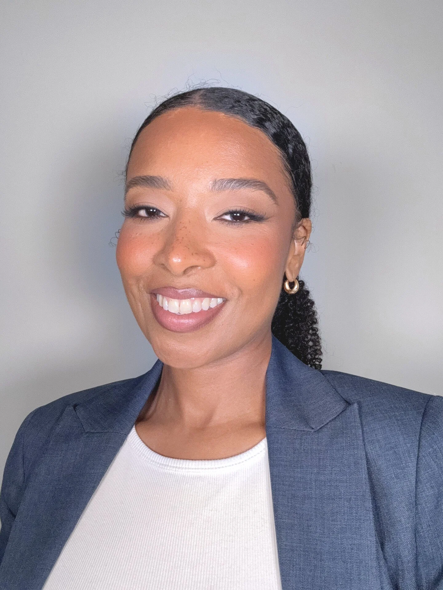 Close-up of a smiling woman with curly hair tied back, wearing a gray blazer and white top, against a plain light background.
