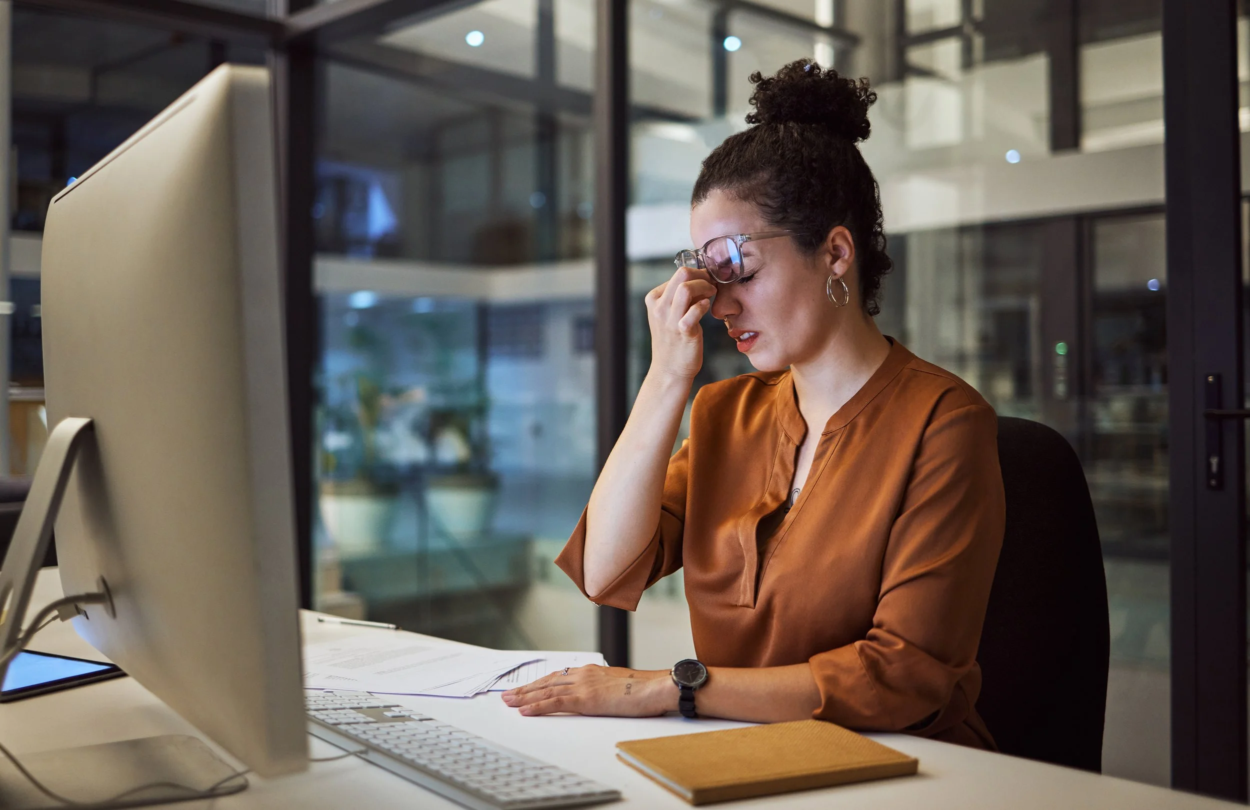 A woman sitting at a desk in an office, looking distressed, holding her glasses with one hand and resting her head on the other, with papers and a computer monitor in front of her.