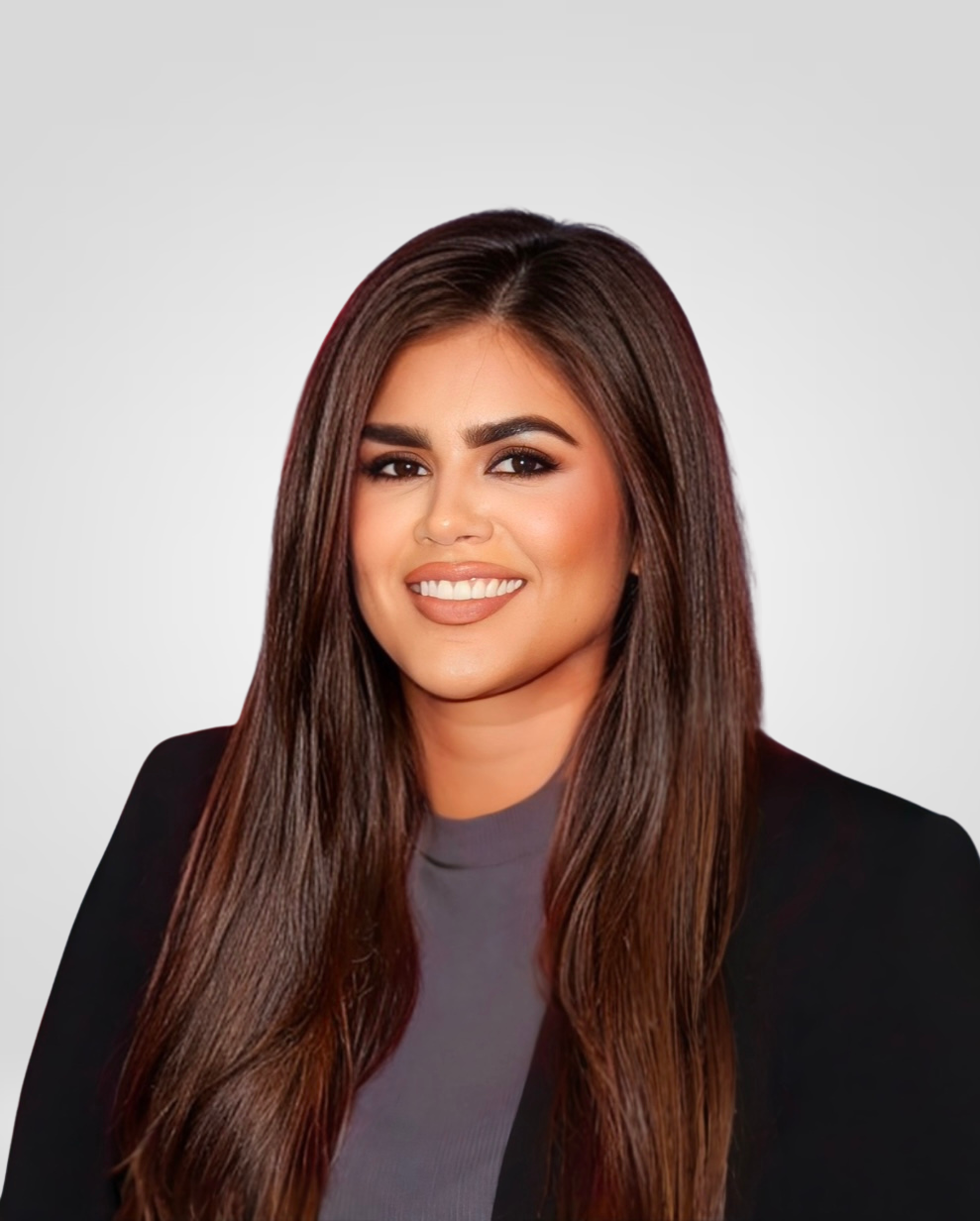 A woman with long brown hair and light makeup, smiling, wearing a black blazer and gray top, against a plain light gray background.