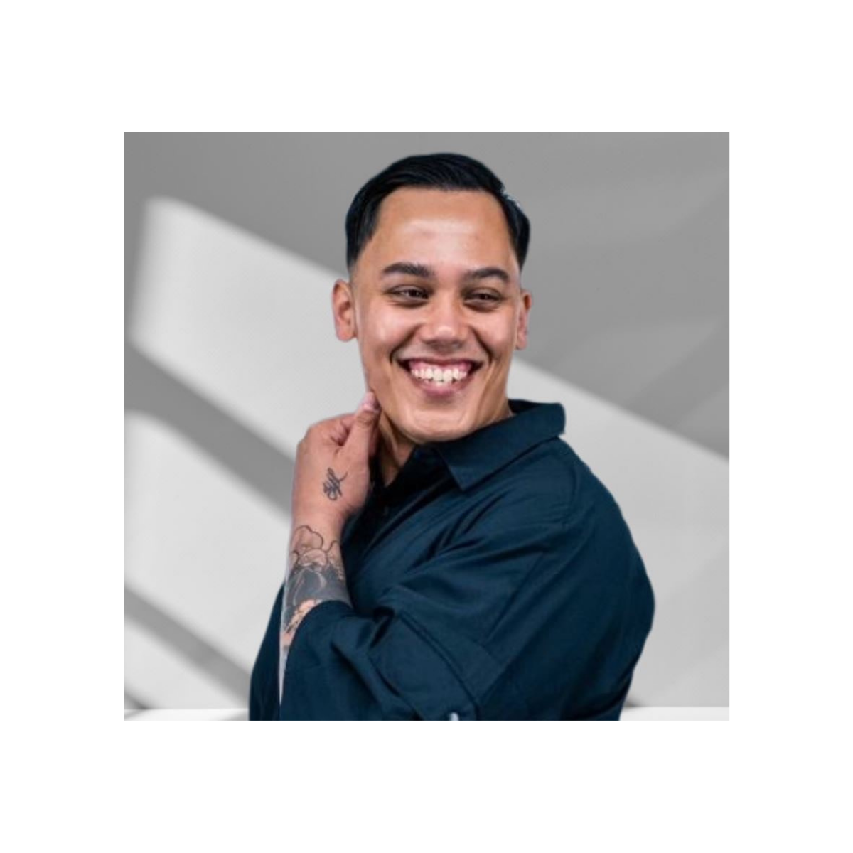 Young man with dark hair, smiling, wearing a dark blue shirt, posing with hand near neck, tattoos on arm, in front of geometric gray and white background.