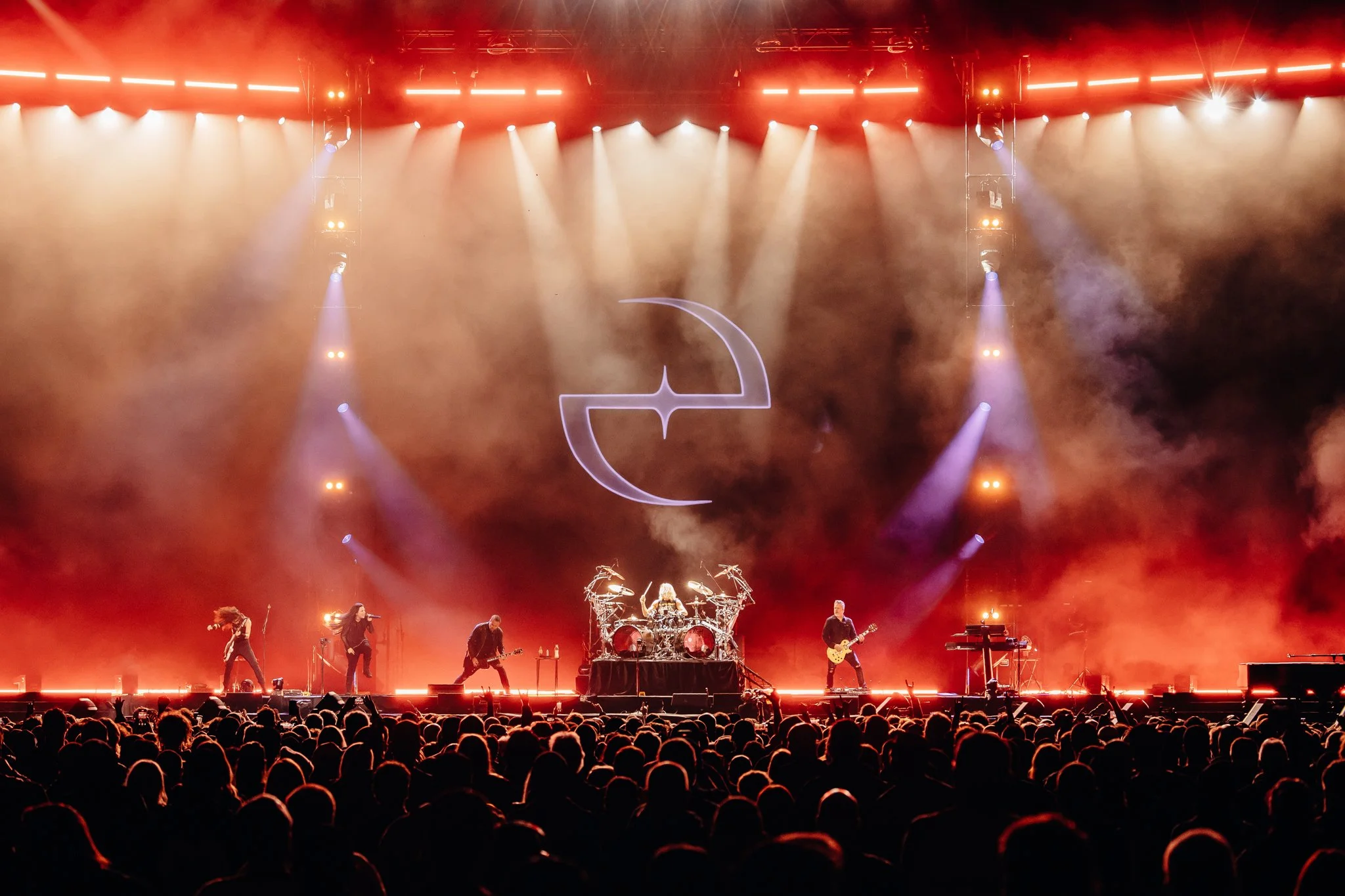 A live concert on a stage with bright lights and a large audience. The stage features a band performing, with a drummer in the center, guitarists on either side, and musical equipment. A large illuminated symbol resembling a crescent moon and star is projected above the performers, and the background has an orange and purple haze from stage lighting.