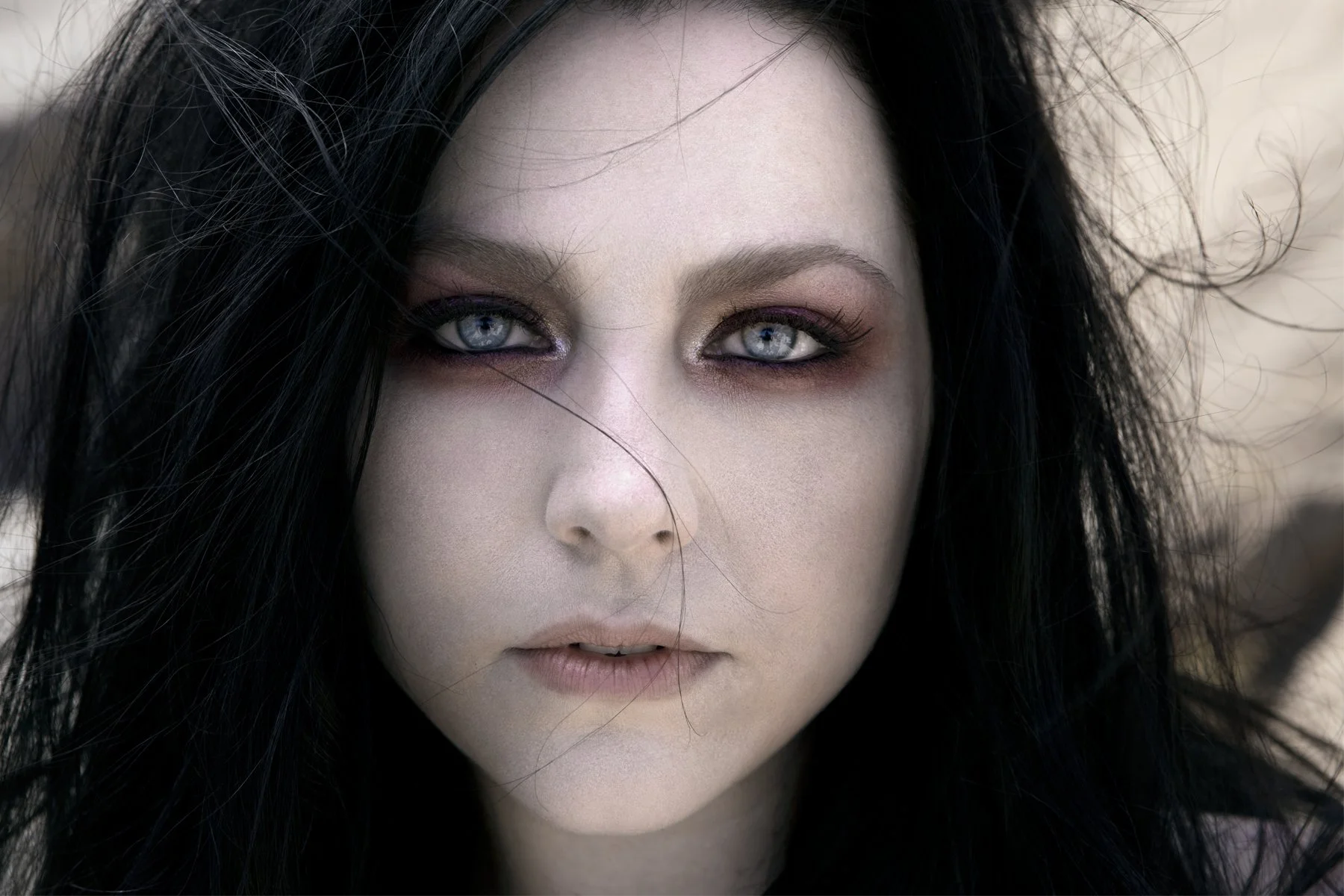 Close-up of a woman's face with piercing blue eyes, pale skin, long dark hair, and makeup with dark eyeshadow and eyeliner.