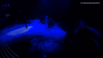 Dark outdoor scene illuminated with blue lights, with fog or smoke on the ground, and a few figures standing in the distance.