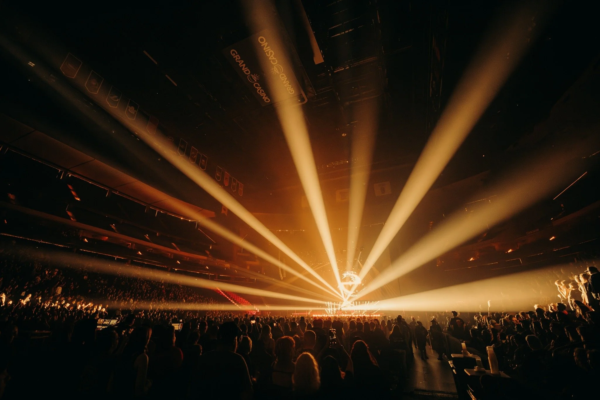 An indoor concert venue with a crowd watching a stage illuminated by yellow spotlights and beams radiating outward, with banners hanging from the ceiling, including one that says 'Grand Casino'.